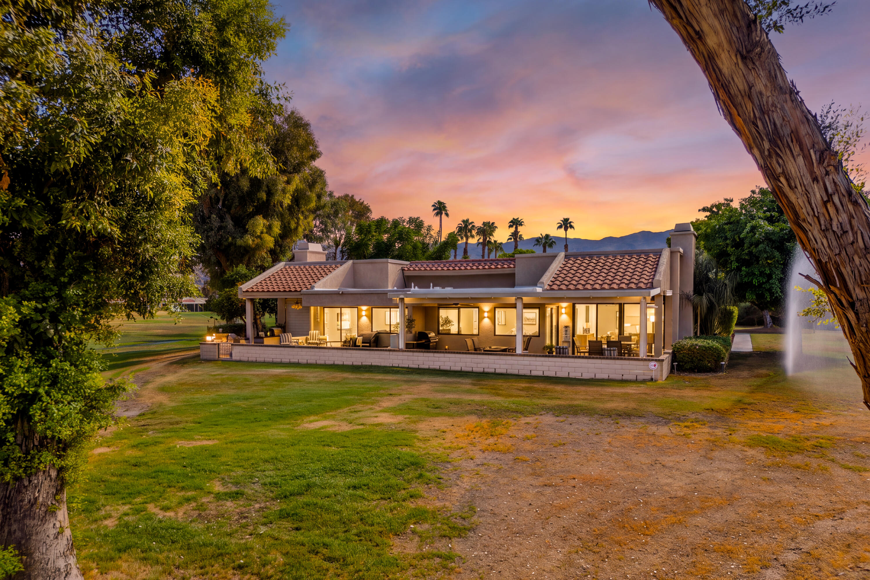 Single-story home with a furnished covered patio, mature trees, and mountains at sunset.