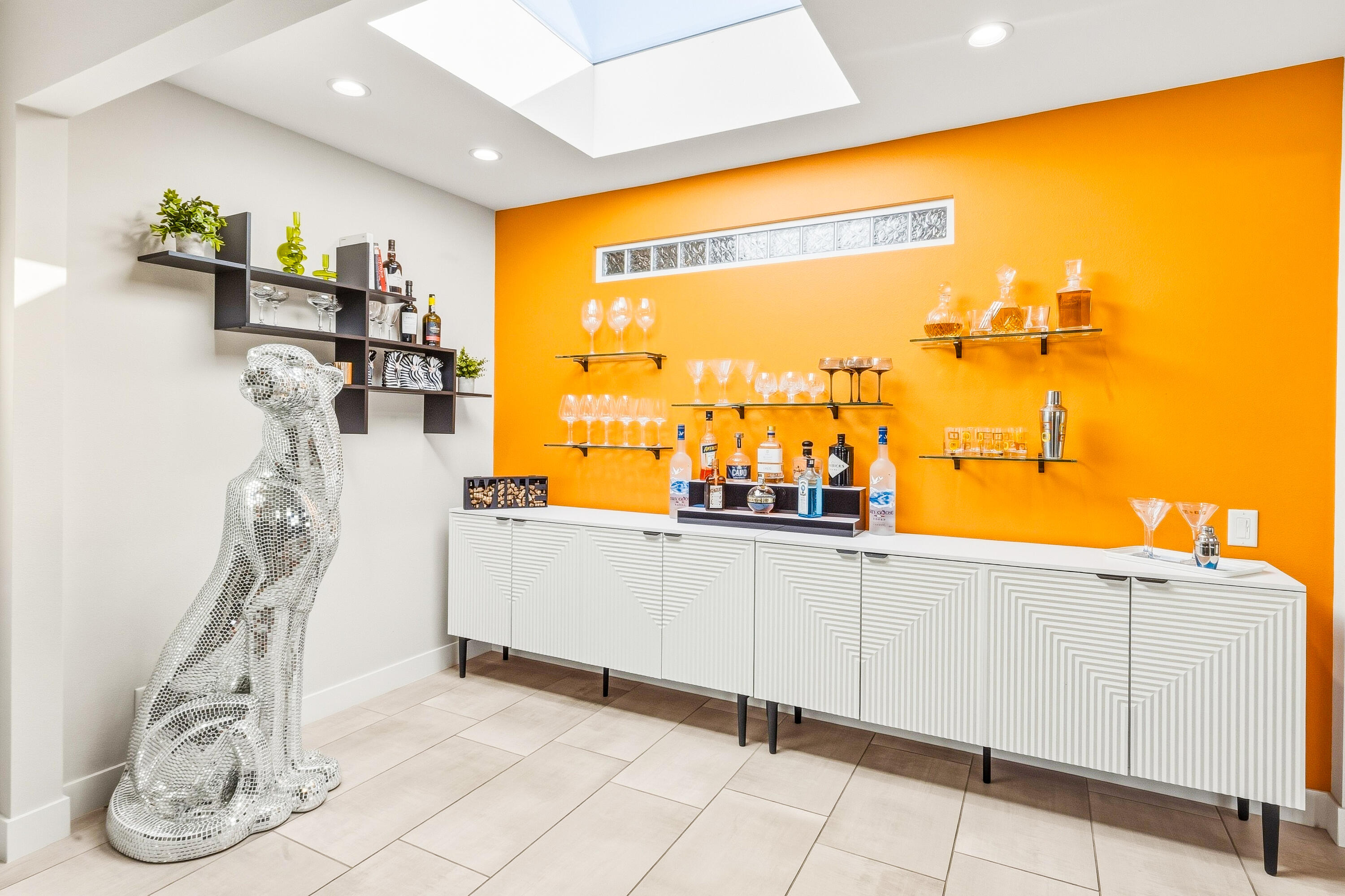 Home bar with orange accent wall, white geometric cabinet, glass shelves, and silver mosaic dog sculpture