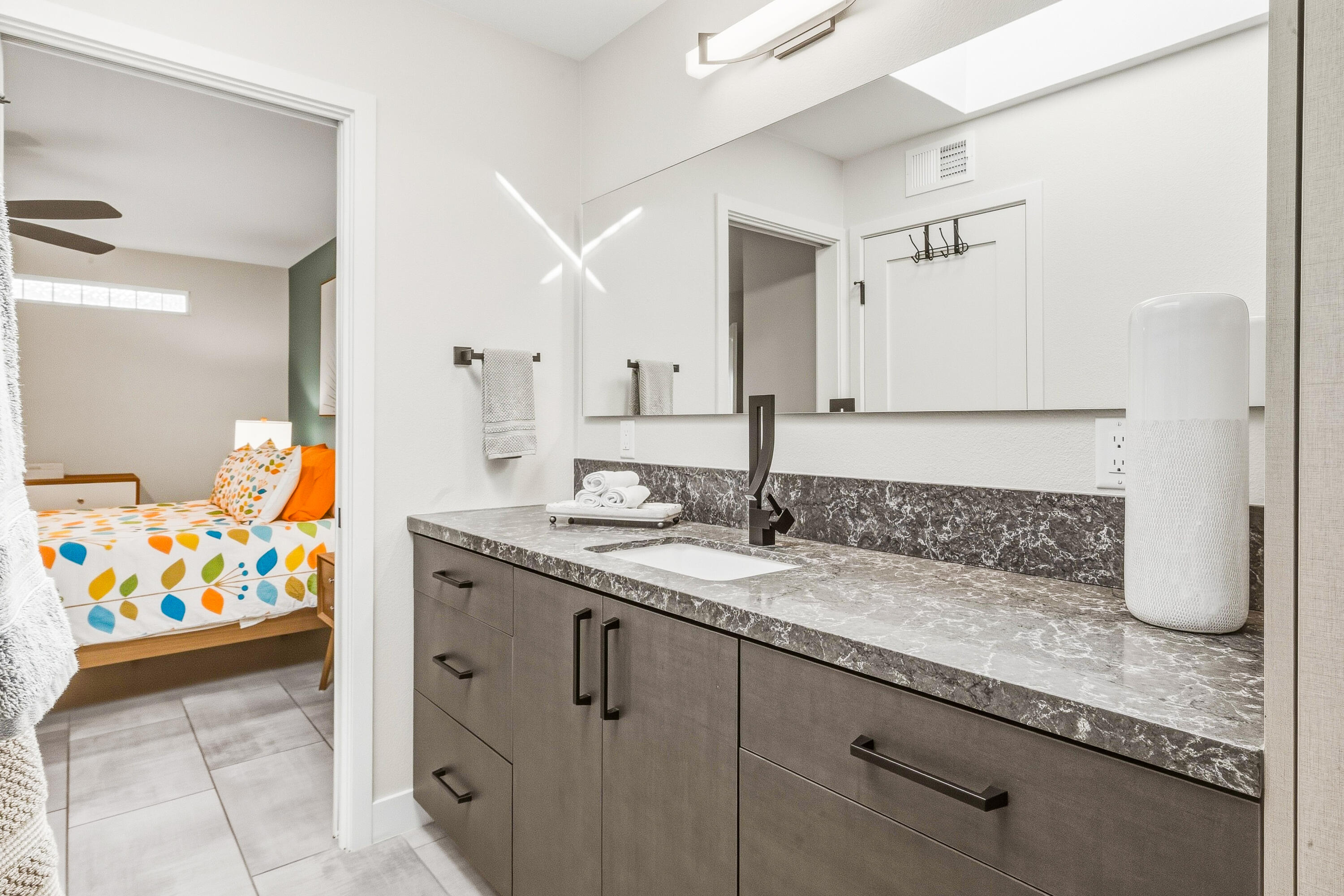 Bathroom vanity with a wide mirror, gray stone-look countertop, and bedroom visible through the doorway.
