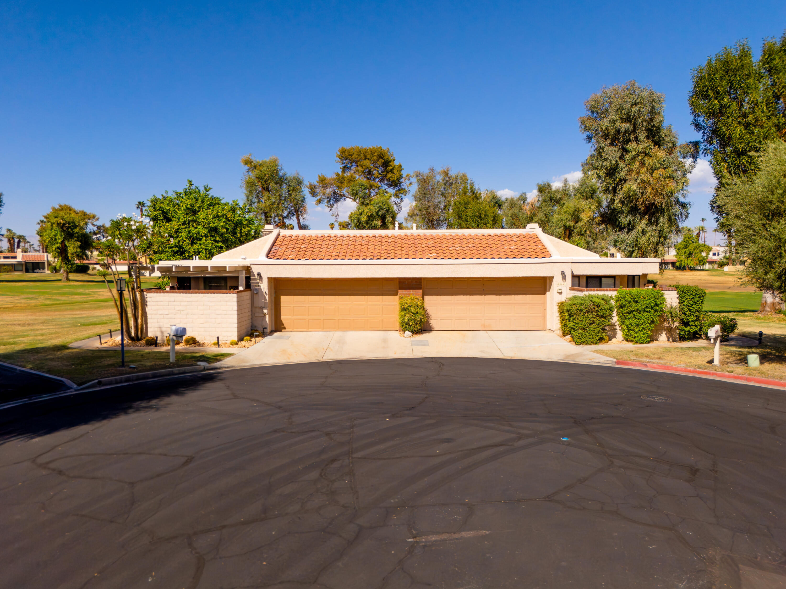 Single-story beige home with a terracotta tile roof, double garage, and shrubs along the driveway.