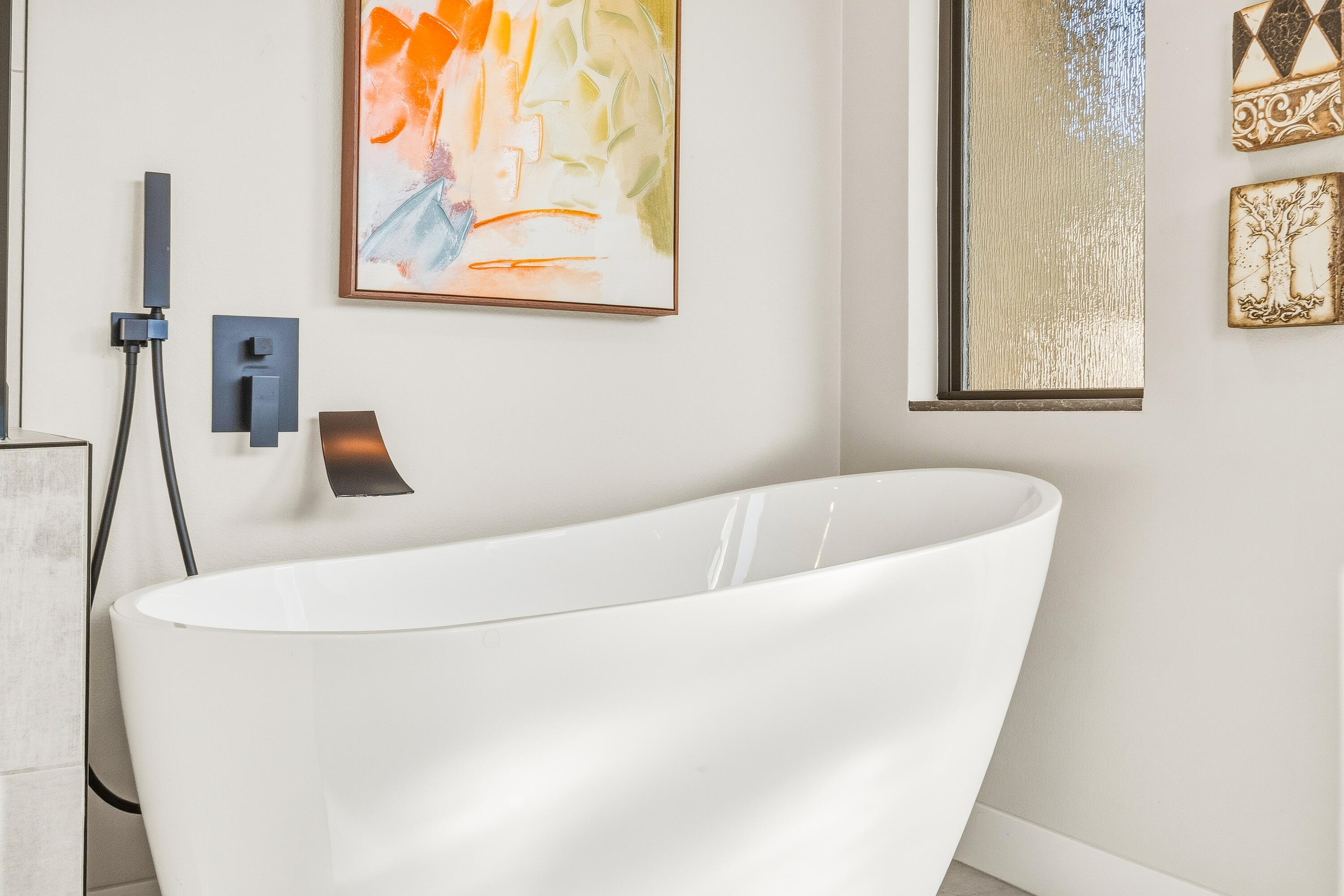 Freestanding white soaking tub with dark wall-mounted fixtures and colorful abstract artwork.