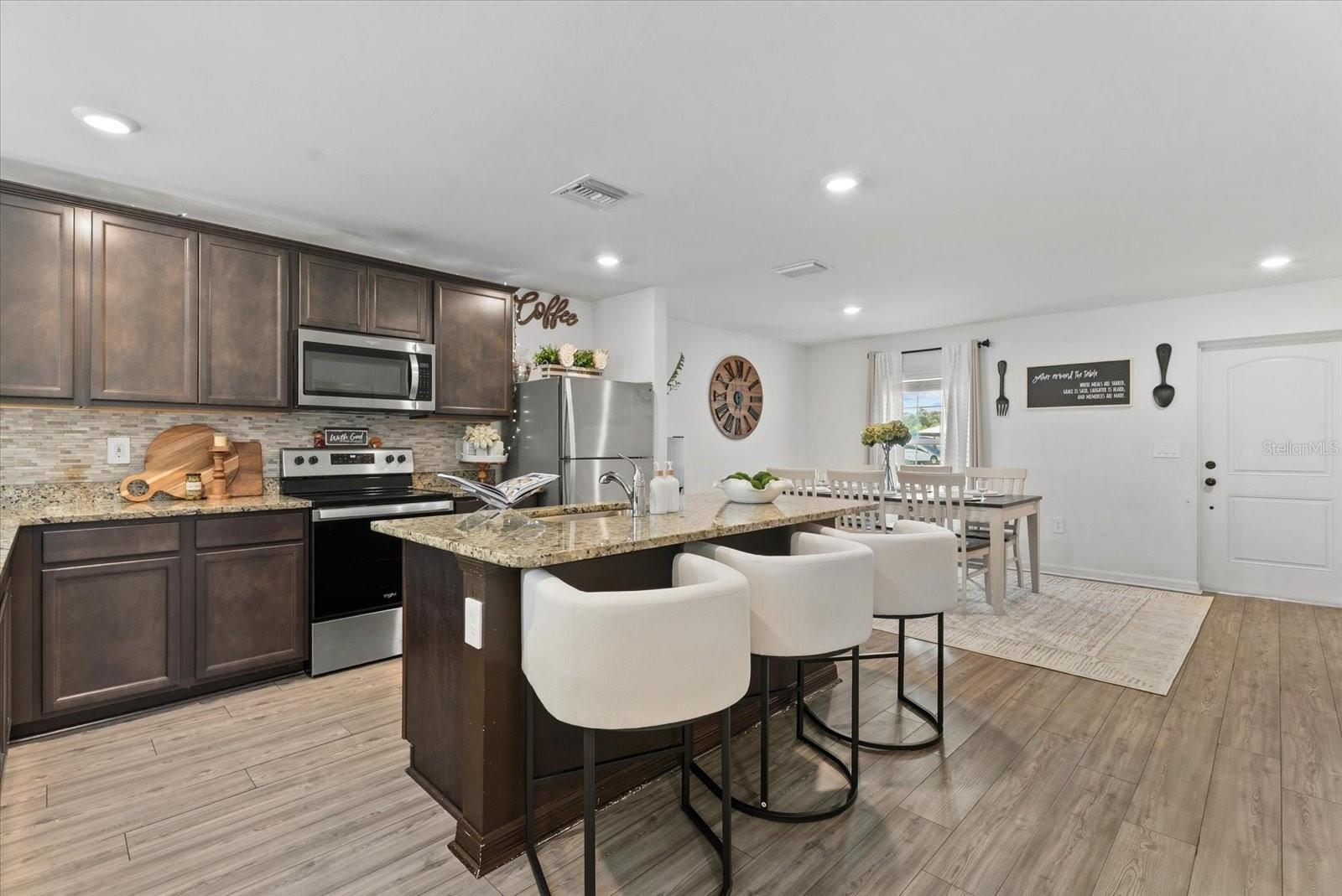 Open-plan kitchen with dark cabinets, granite island, white bar stools, and adjoining dining area.