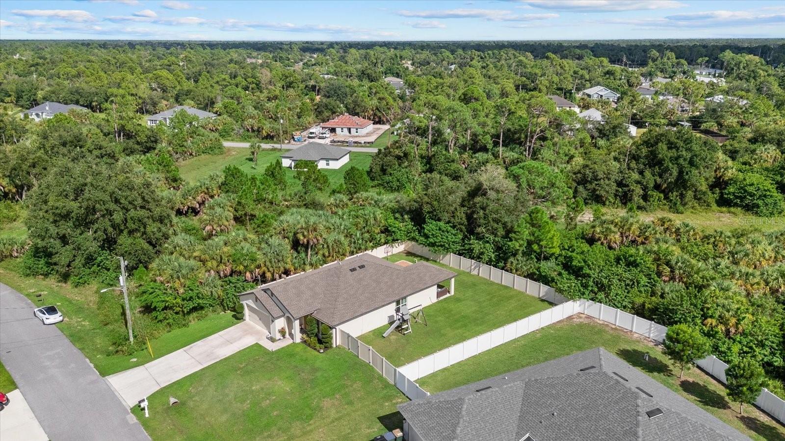 Aerial view of a single-story home with a large fenced lawn and swing set, surrounded by trees.