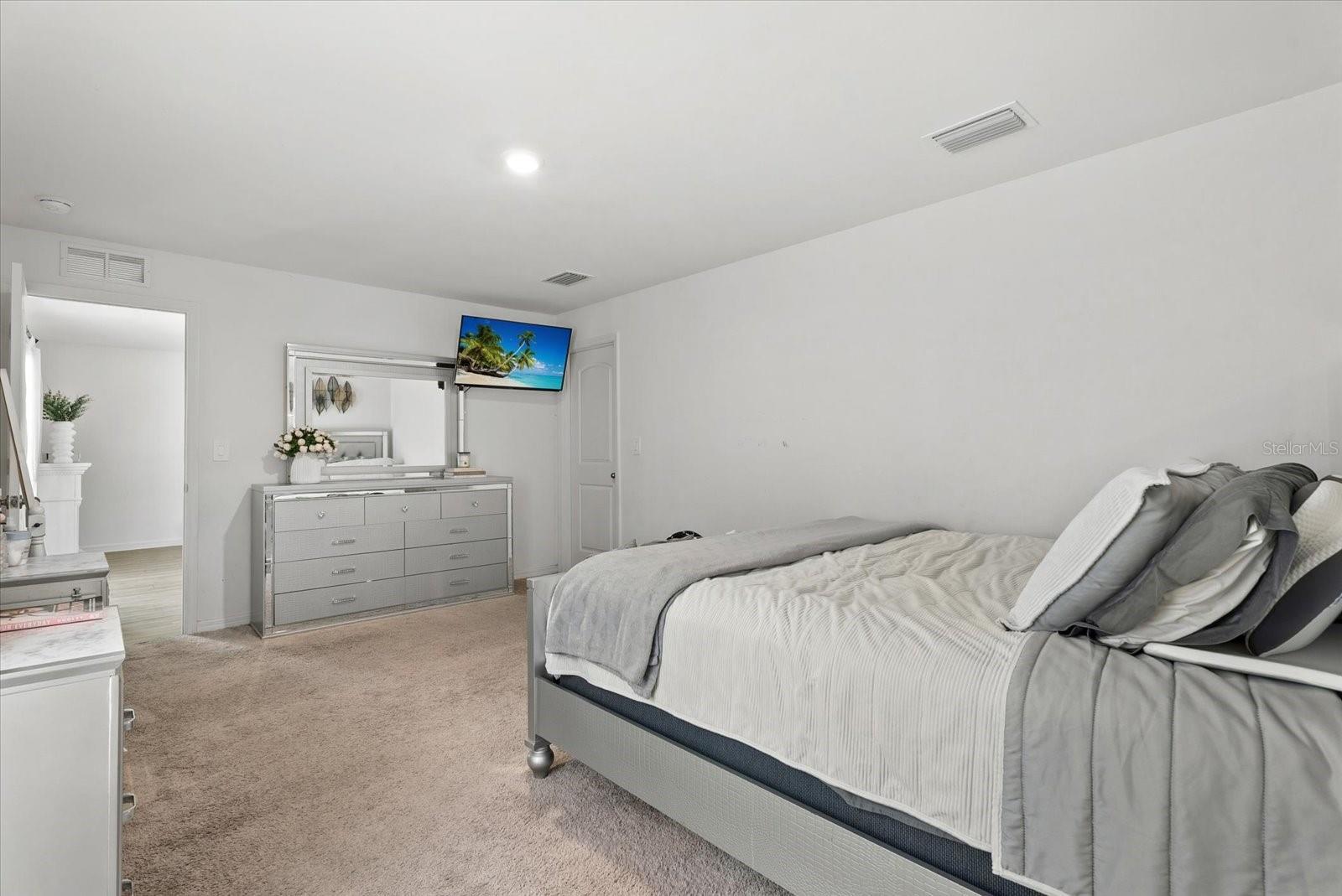 Furnished bedroom with a large gray-and-white bed, mirrored dresser, and wall-mounted television.