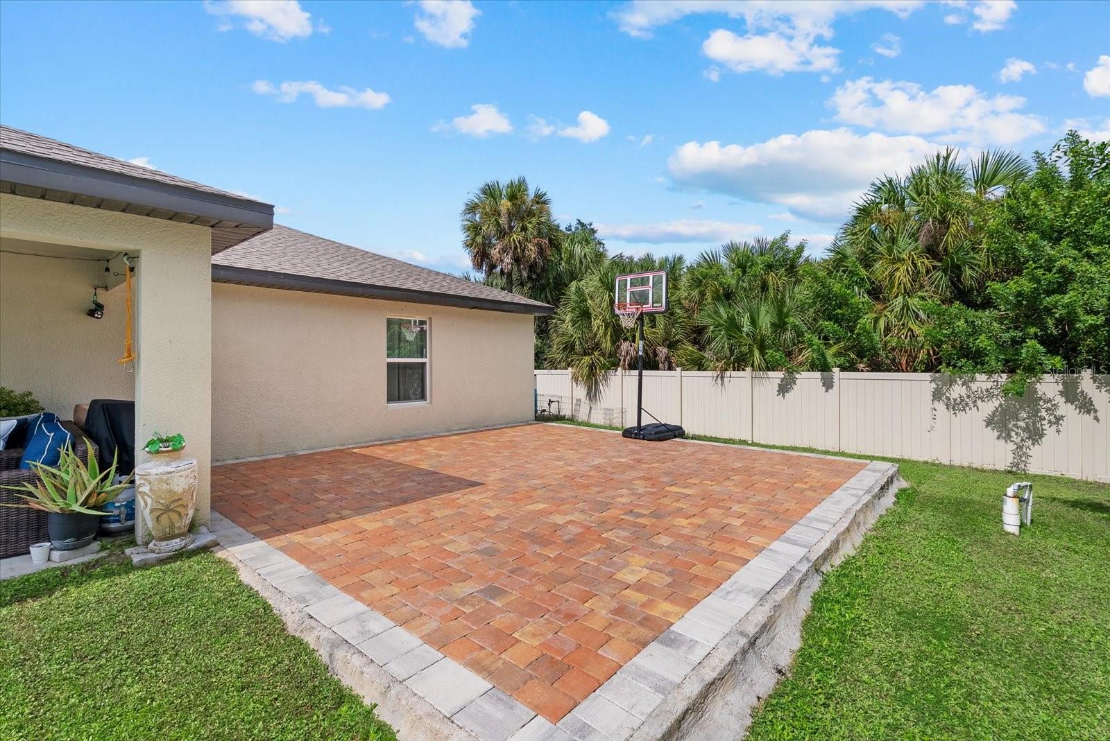 Backyard paver patio with a basketball hoop, covered seating, and palms behind a white privacy fence.