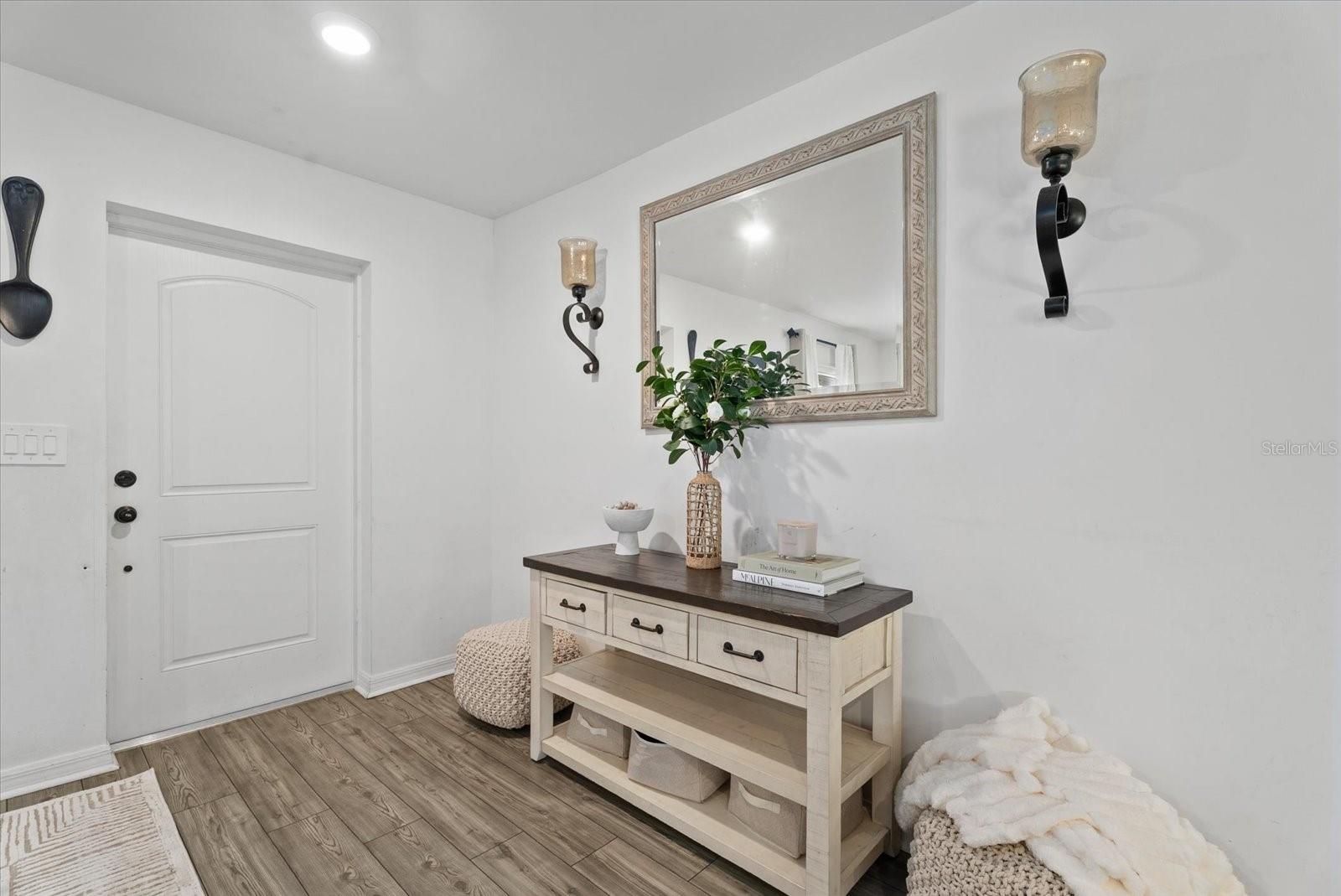 Bright entryway with a white front door, console table, framed mirror, and wood-look flooring.