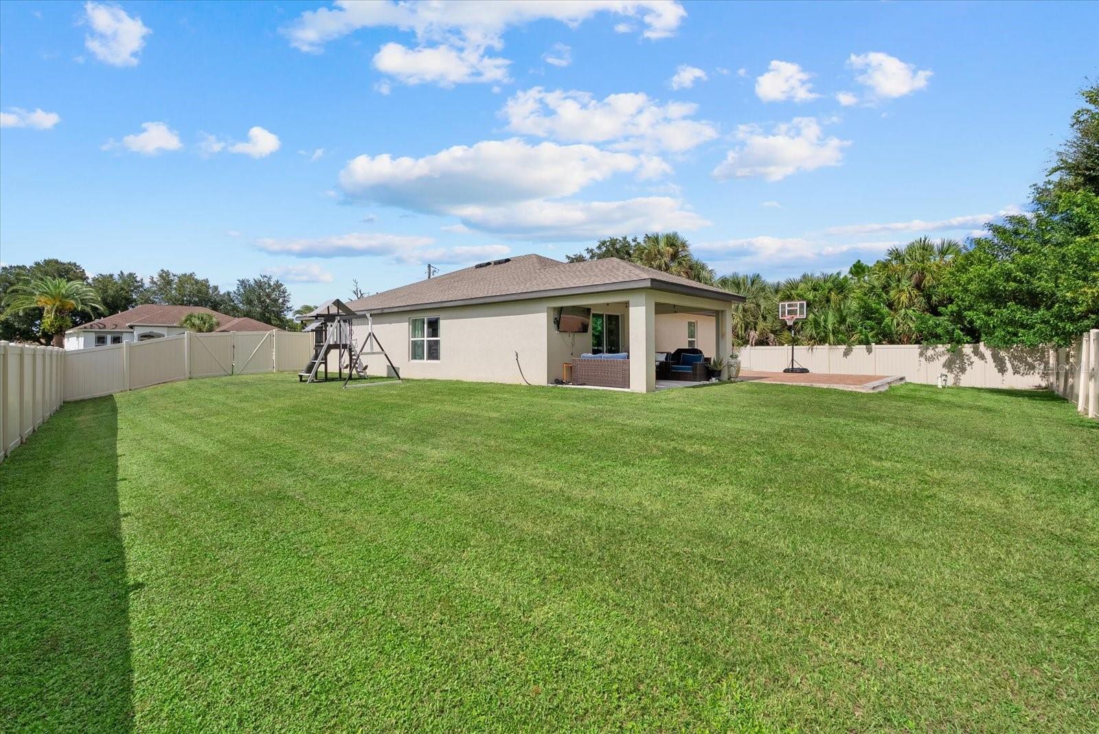 Fenced backyard with broad lawn, covered patio, playset, and basketball hoop.