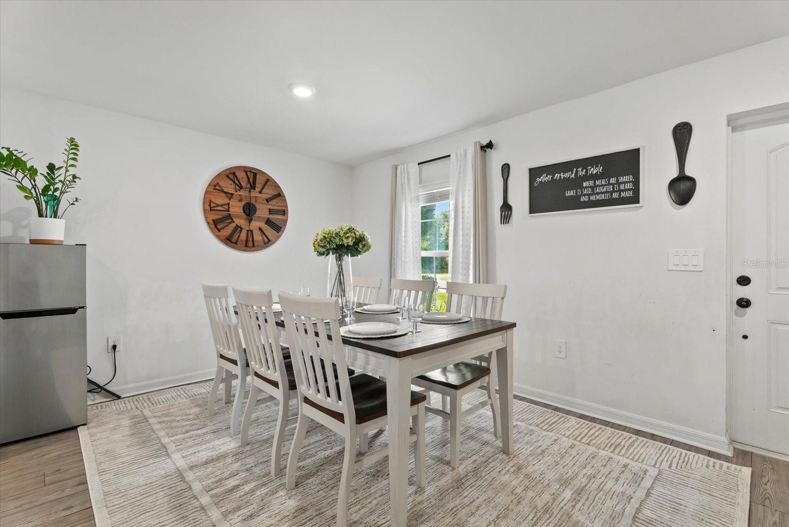 Dining room with a set white table, large wooden wall clock, and window with white curtains.