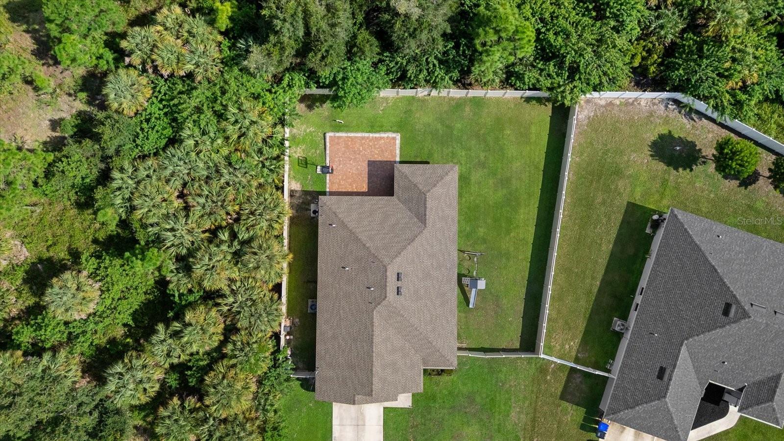 Aerial view of a home with a fenced lawn, paved patio, and dense palms along one side.