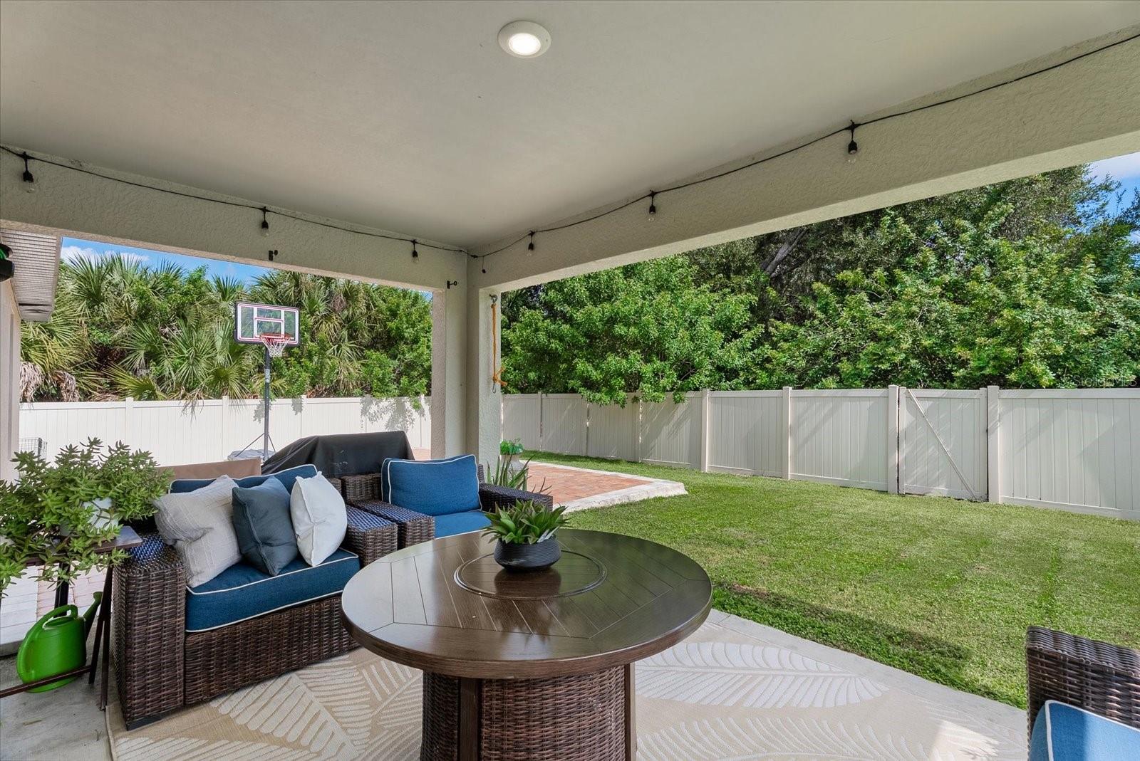 Covered patio with blue wicker seating and a view of the fenced, tree-lined yard.