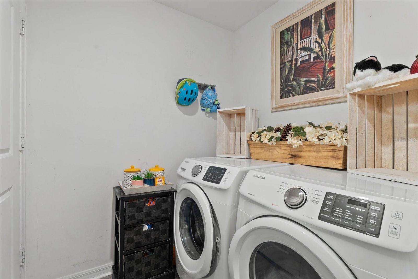 Laundry room with white front-loading washer and dryer and flowers displayed above them.