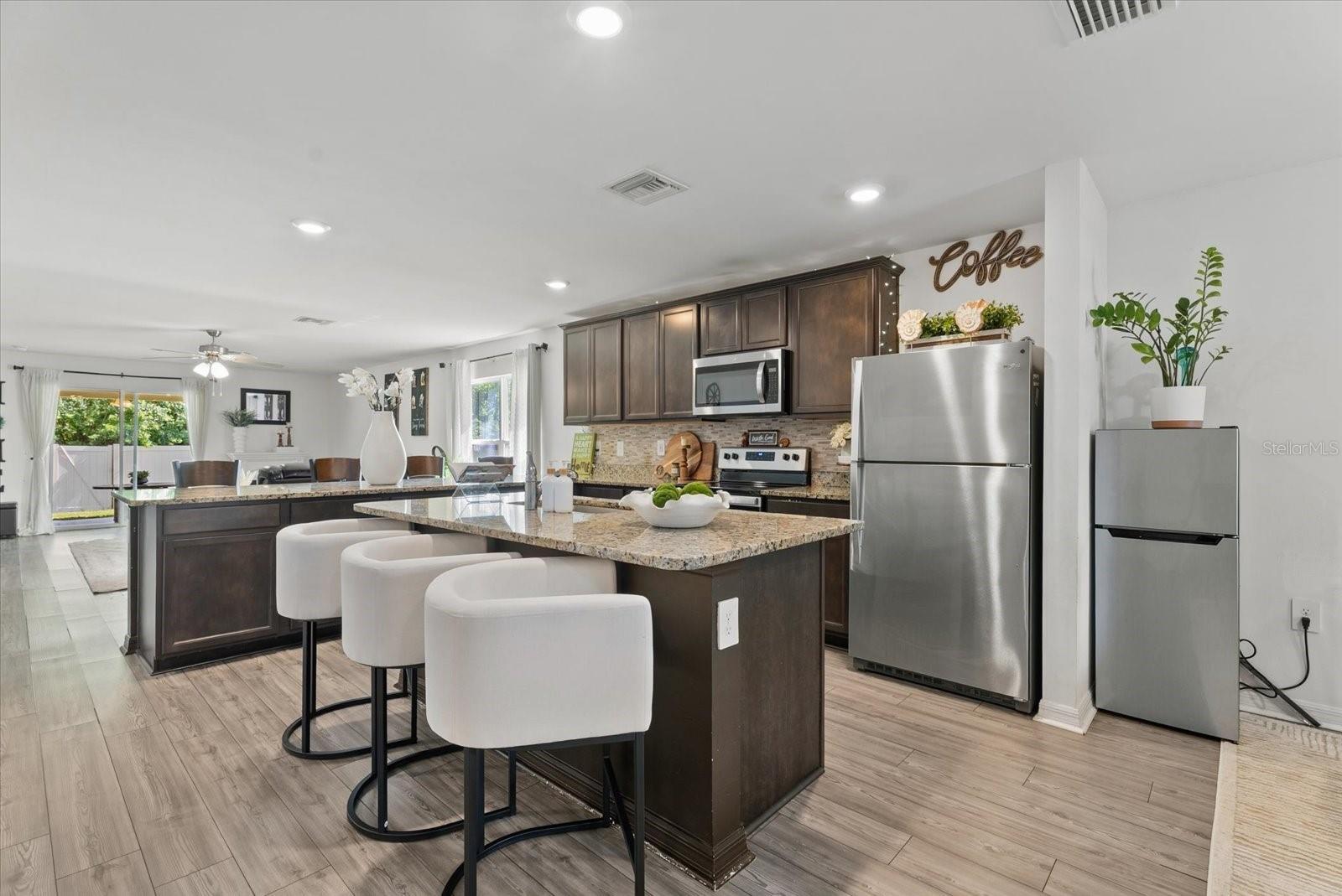 Kitchen with a granite island, white bar stools, dark cabinets, and stainless steel appliances.