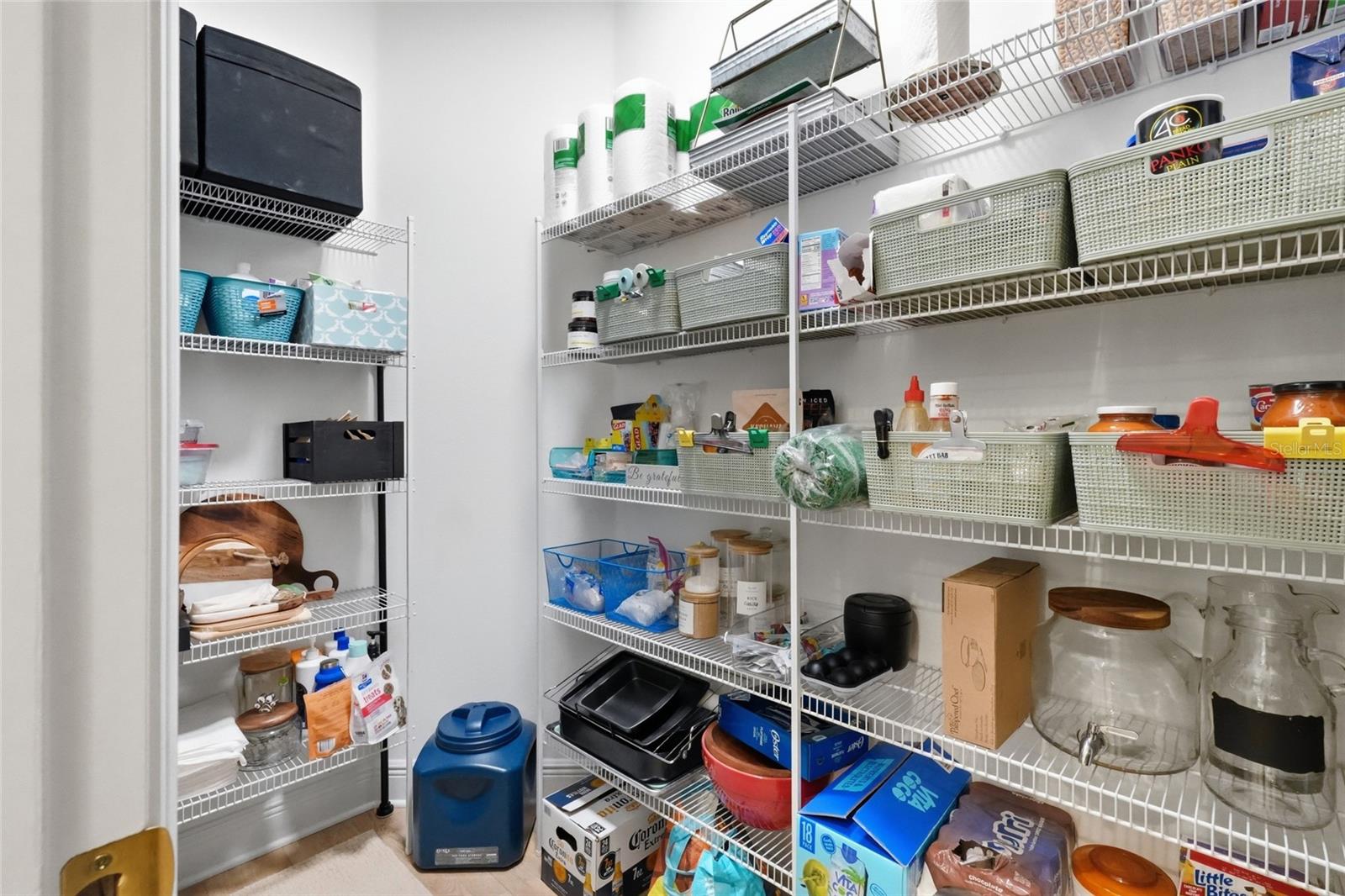 Walk-in pantry with white wire shelving, woven baskets, and neatly stored food and household supplies.