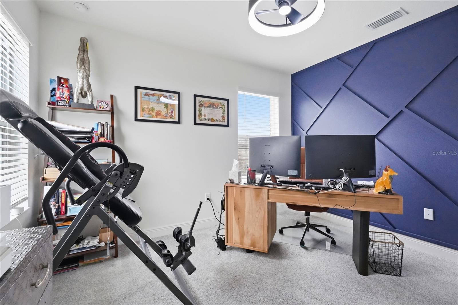 Home office with a blue geometric accent wall, dual-monitor desk, and exercise equipment.