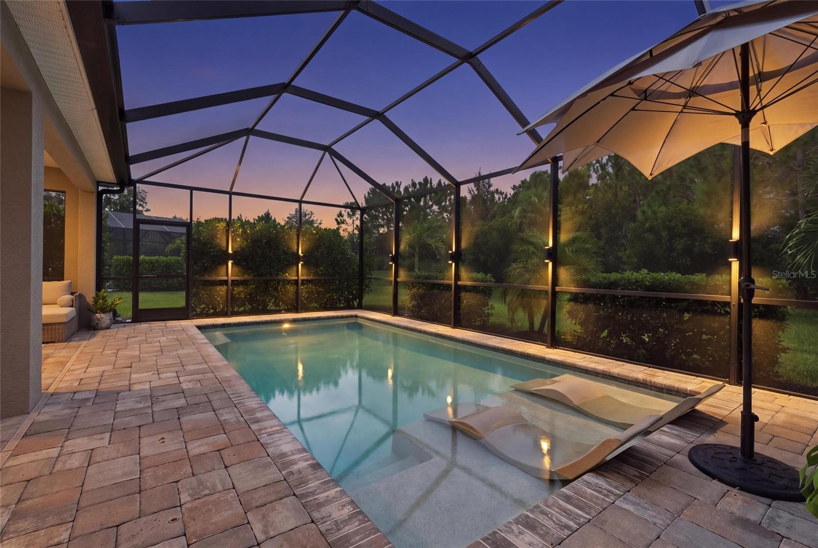 Screened backyard pool at dusk with a shallow sun shelf, in-water loungers, and paver deck.