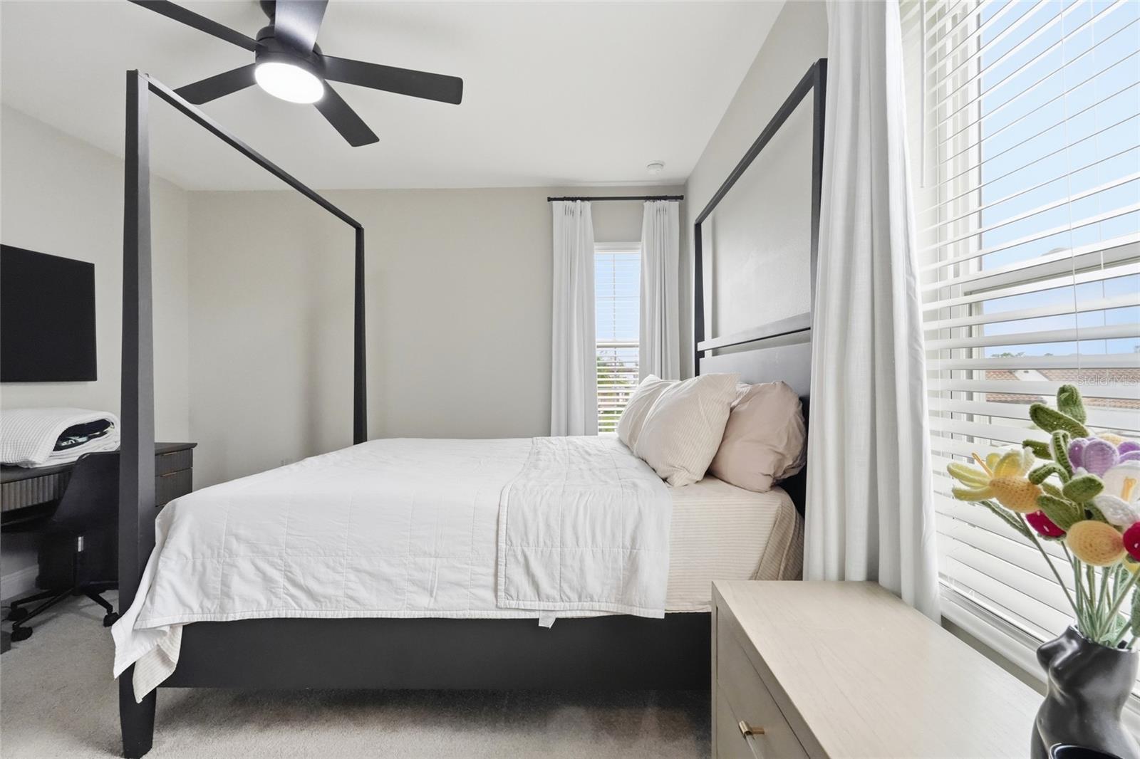 Bright bedroom with a black four-poster bed, white bedding, and large windows with blinds.
