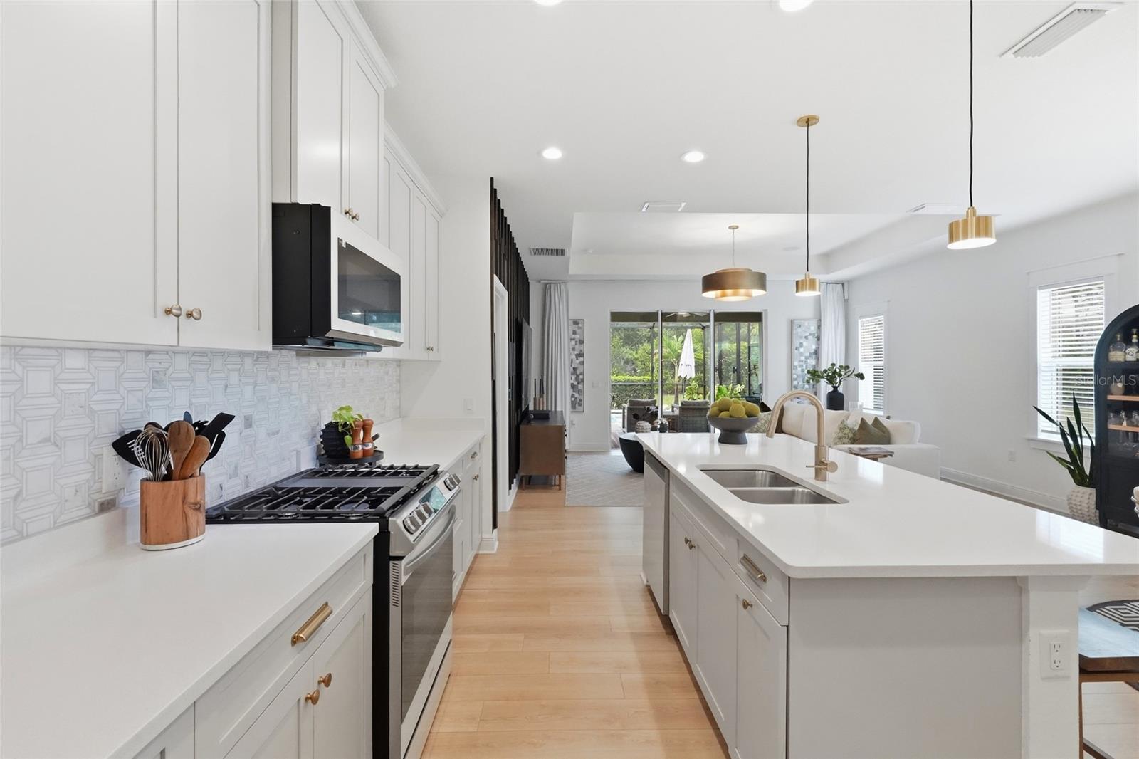 Bright white kitchen with a large island, gold faucet, patterned backsplash, and living area beyond.