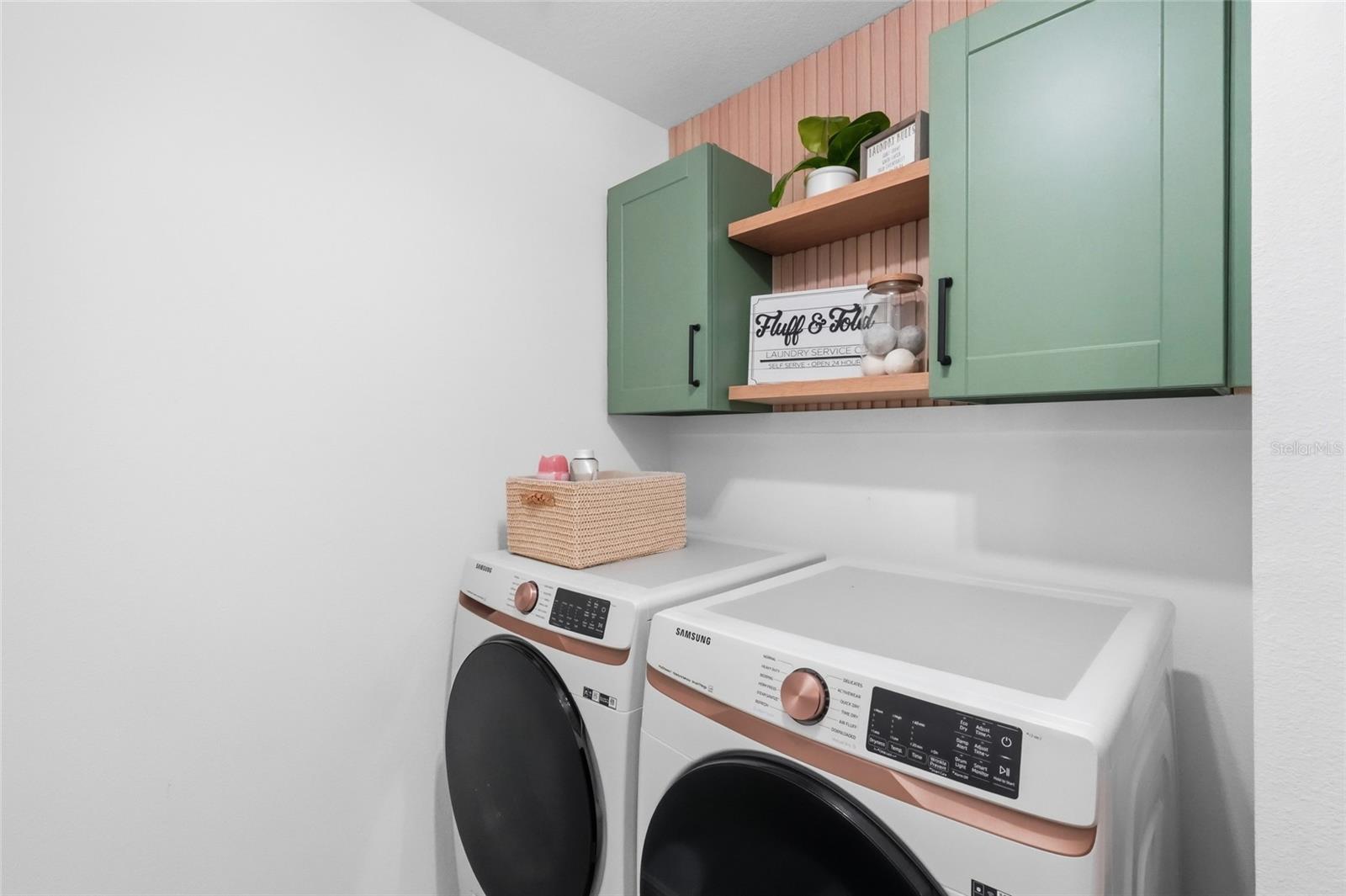 Laundry room with white front-loading washer and dryer beneath green cabinets and a wooden shelf.