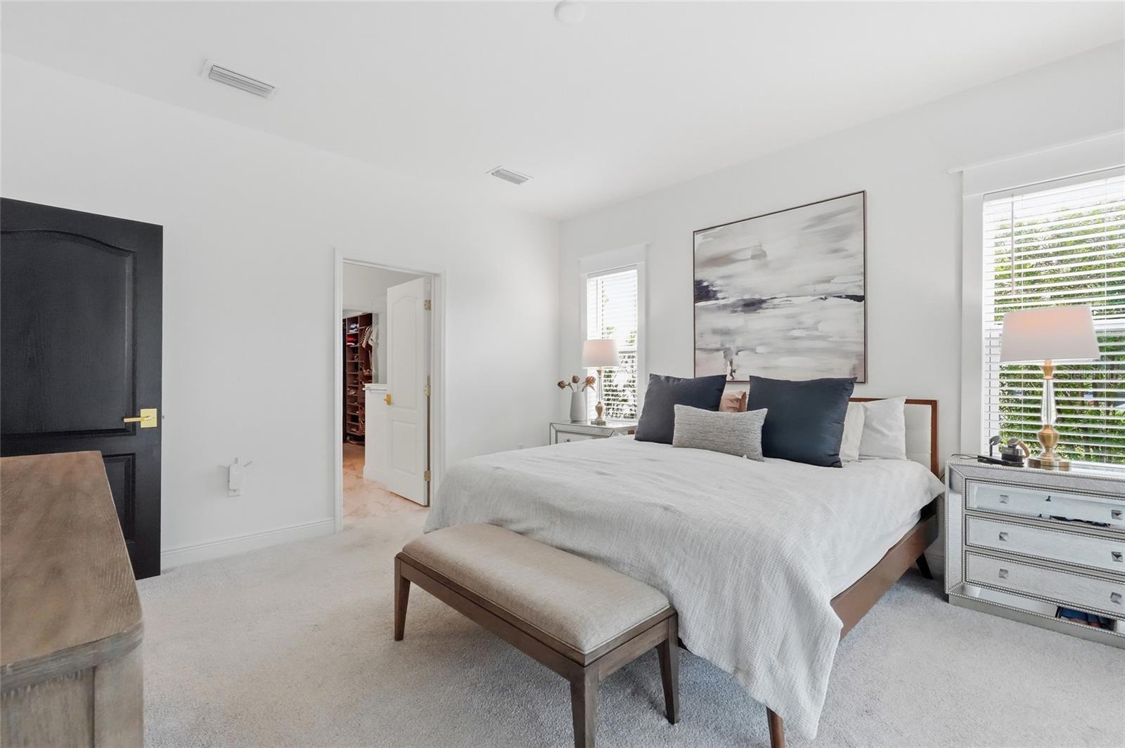 Bright bedroom with a large white bed, dark pillows, cushioned bench, and abstract wall art.