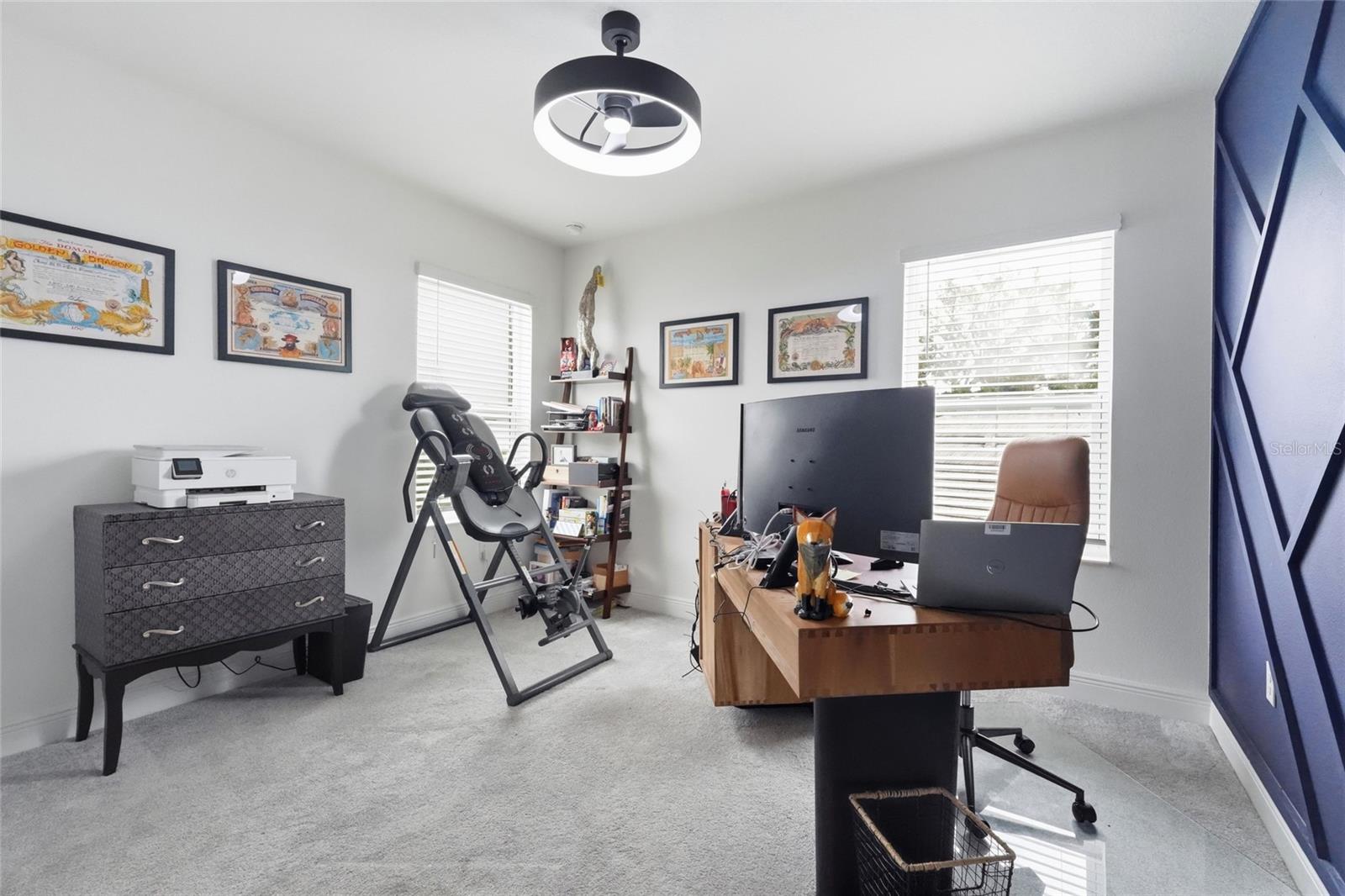 Home office with a wooden desk, computer monitors, an inversion table, and a blue geometric accent wall.