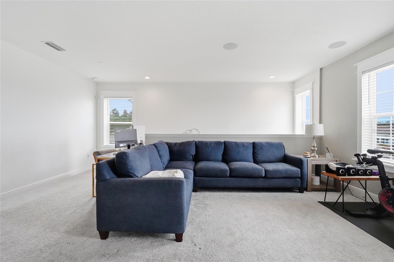 Living room with a navy sectional, light carpeting, bright windows, and exercise equipment.