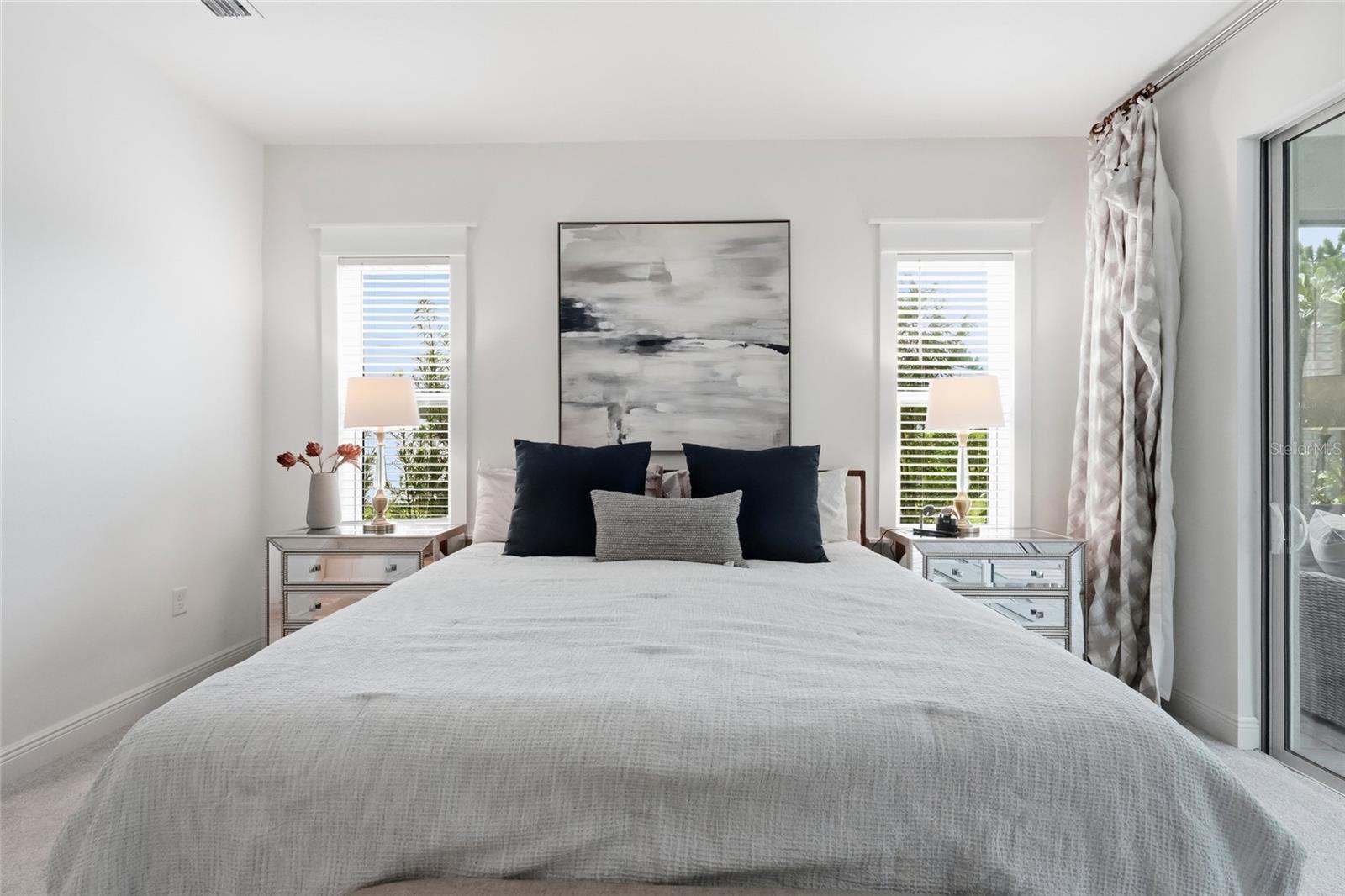 Staged bedroom with a white bed, navy pillows, mirrored nightstands, and a sliding glass door.