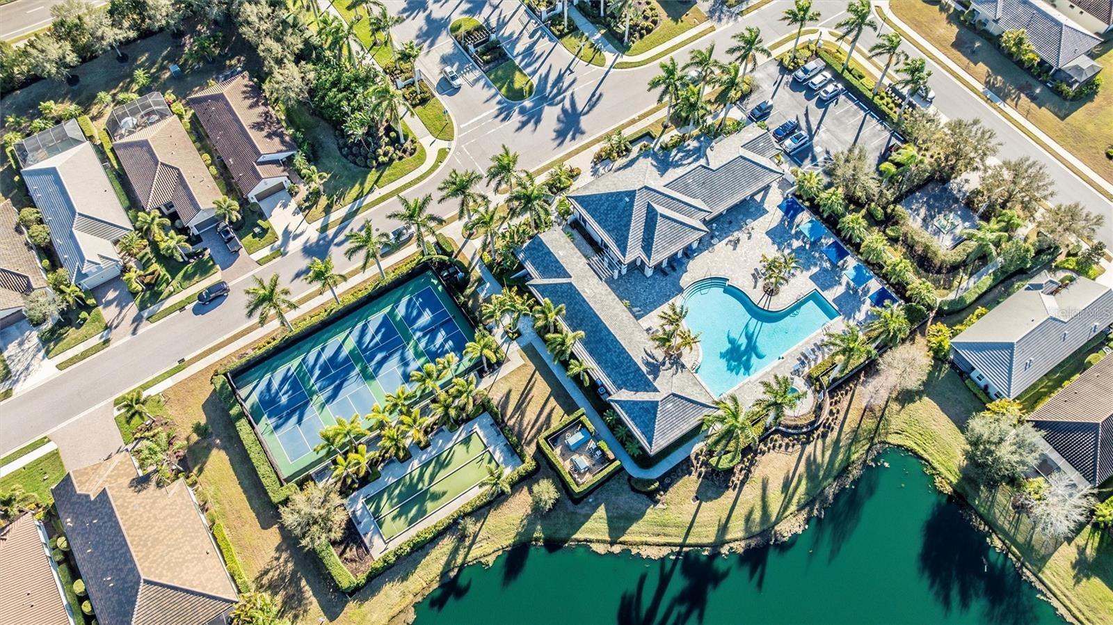 Aerial view of a home beside a pool and tennis courts, with a pond and palm-lined neighborhood.