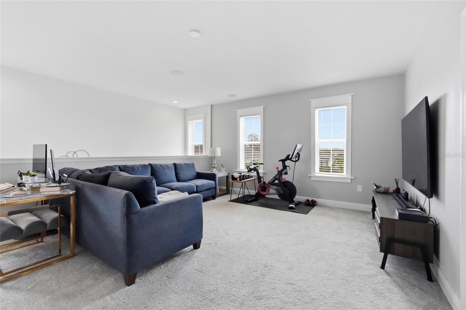 Living room with a blue sectional, desk, television, and exercise bike beside three tall windows.