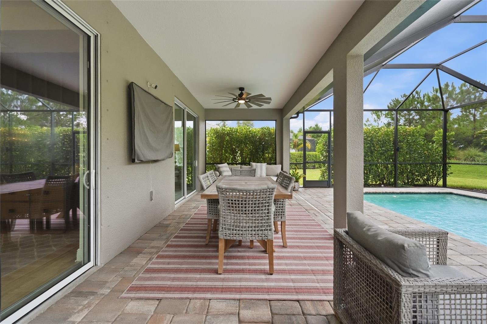 Screened lanai with wicker dining furniture beside a pool and leafy hedges.