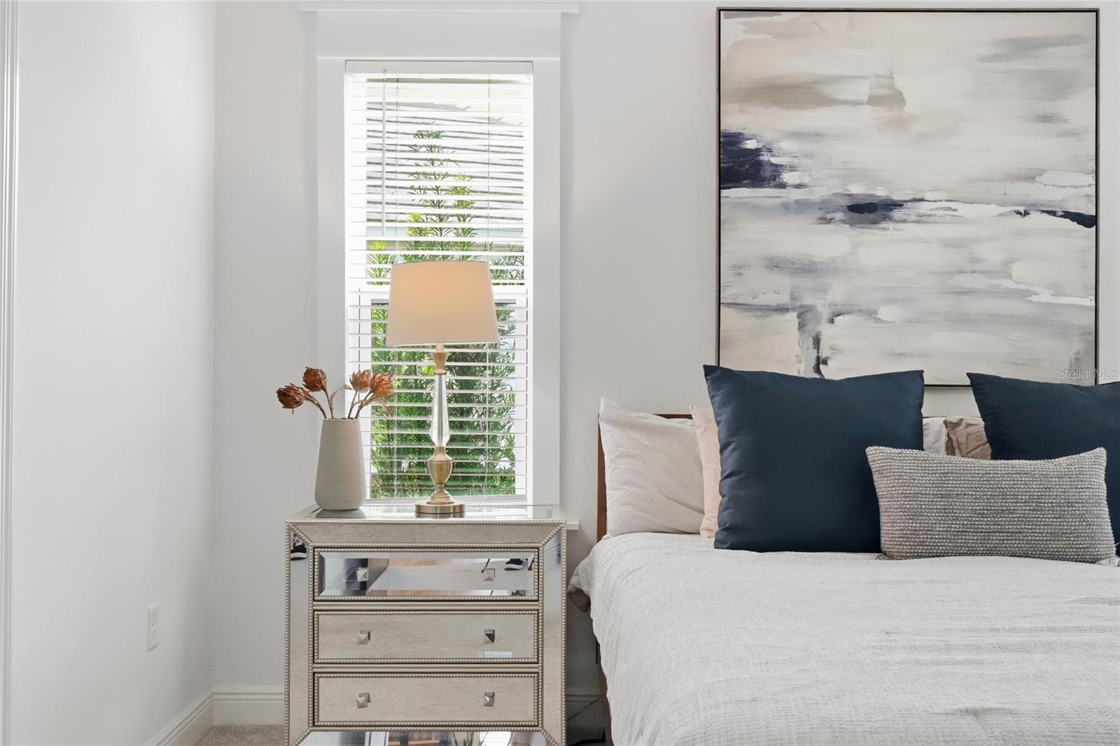 Bedroom with a white bed, navy pillows, mirrored nightstand, and large abstract painting.