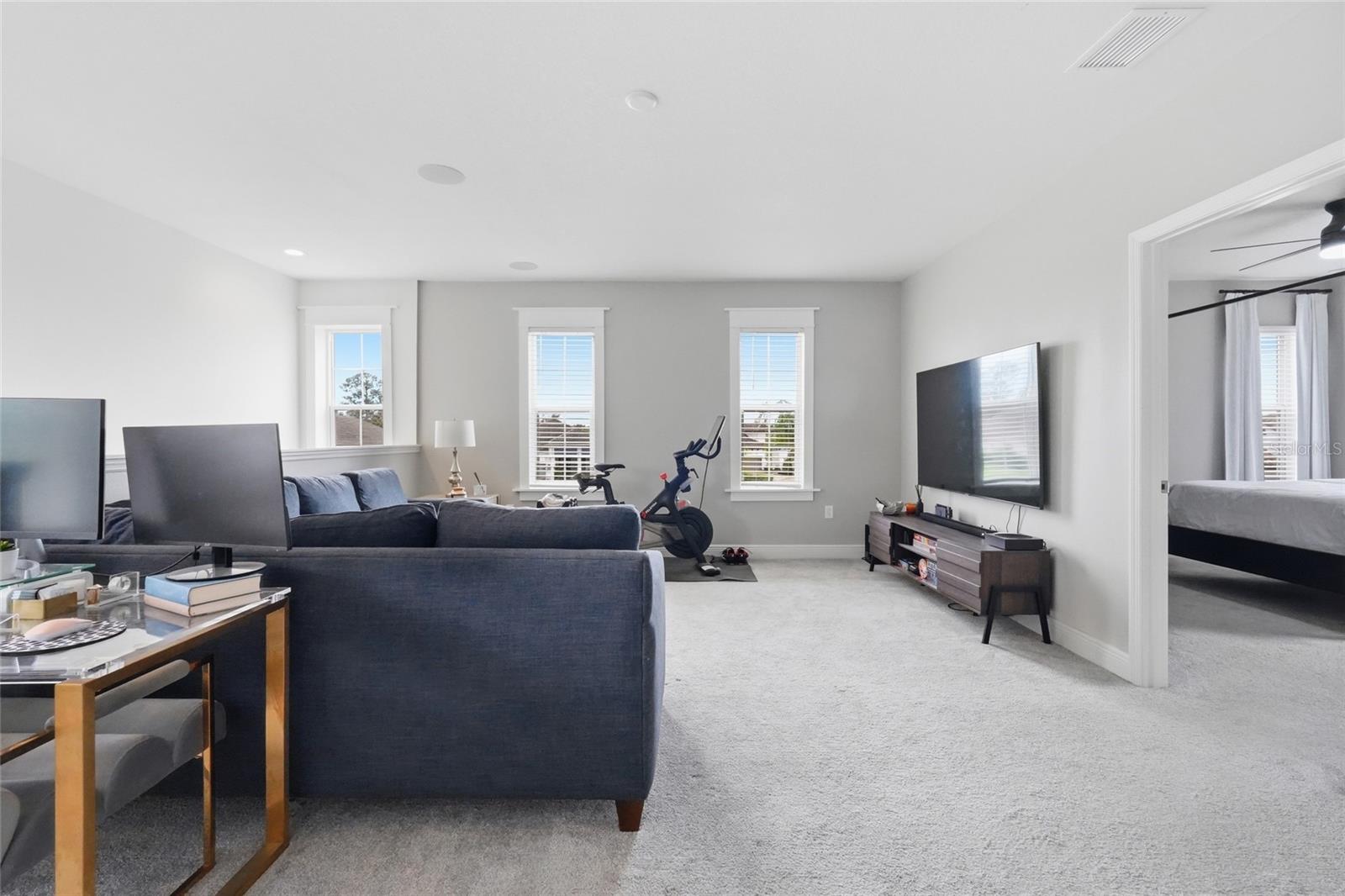 Furnished living room with navy sofas, a wall-mounted TV, and an exercise bike by tall windows.