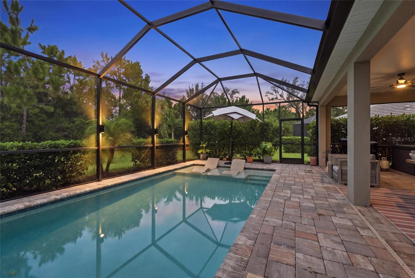 Screened backyard pool at dusk with in-water loungers and a paved patio beside the house.