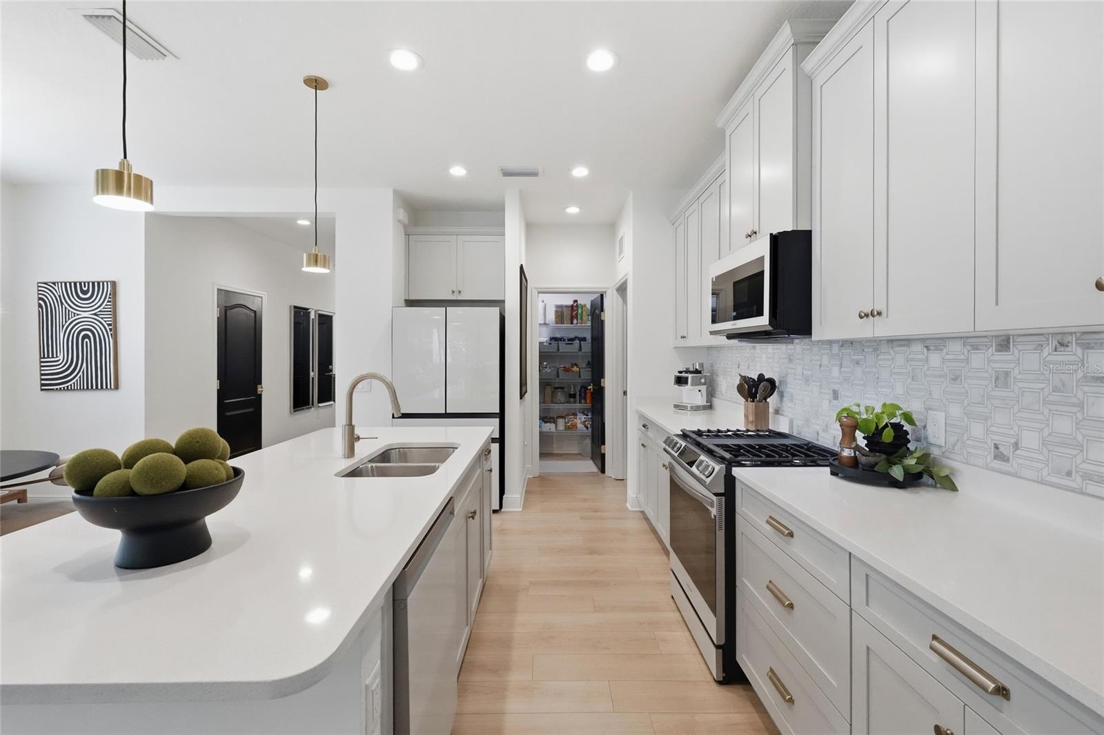 Modern white kitchen with a long sink island, patterned backsplash, stainless appliances, and open pantry.