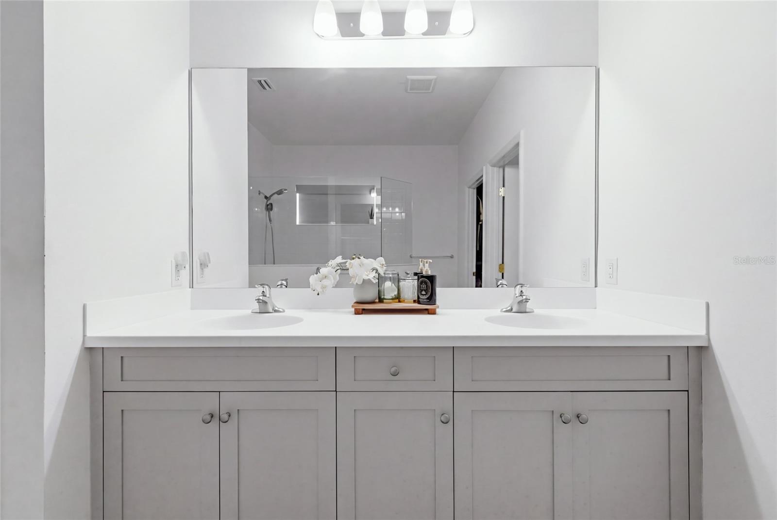 Bathroom with a white double-sink vanity, gray cabinets, and a large mirror reflecting the glass shower.