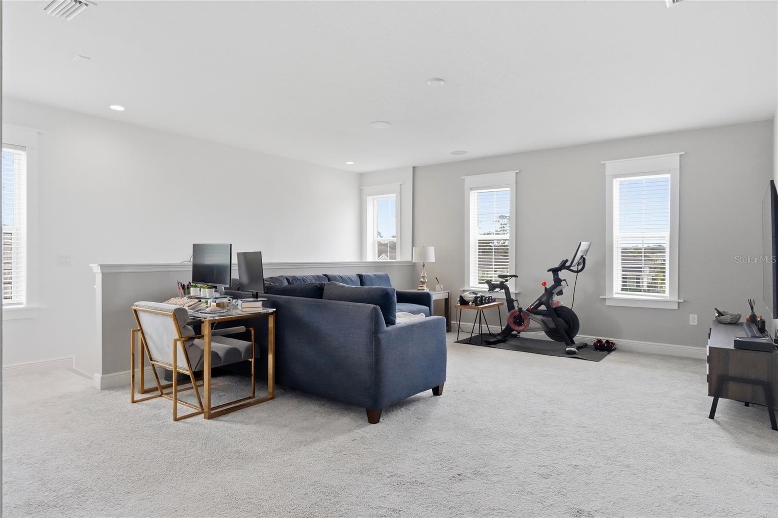 Furnished living area with a blue sectional, desk, exercise bike, and three tall windows.
