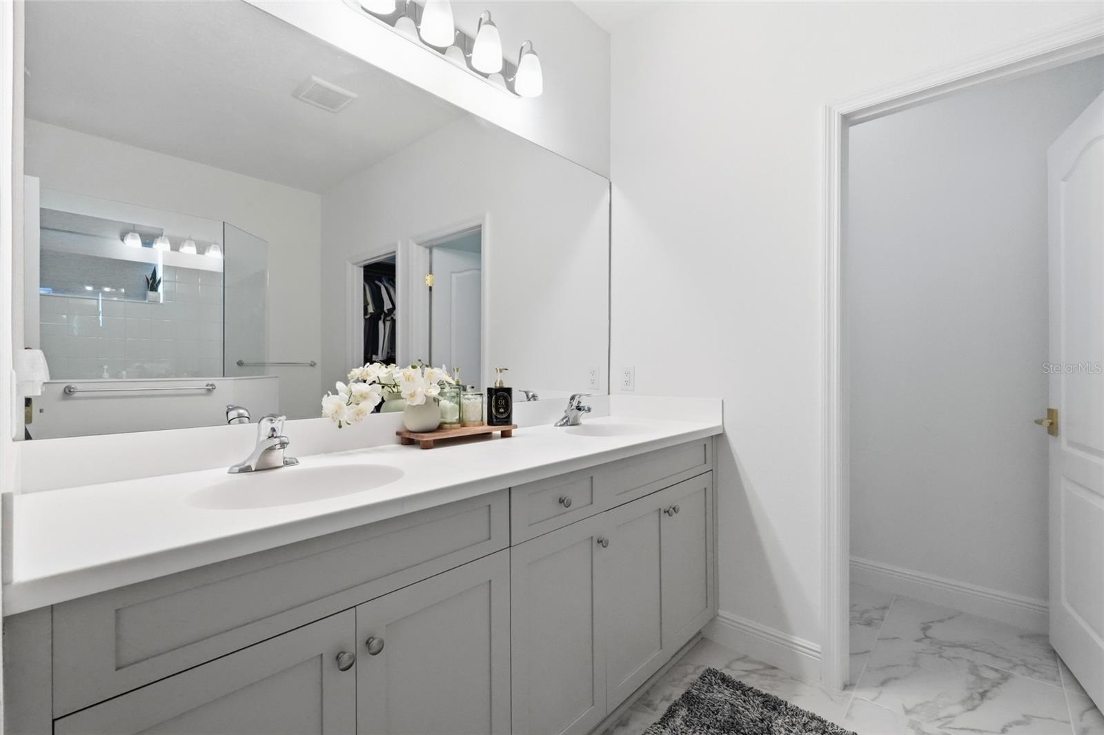 Bathroom vanity with double sinks, gray cabinetry, a large mirror, and flowers on the countertop.