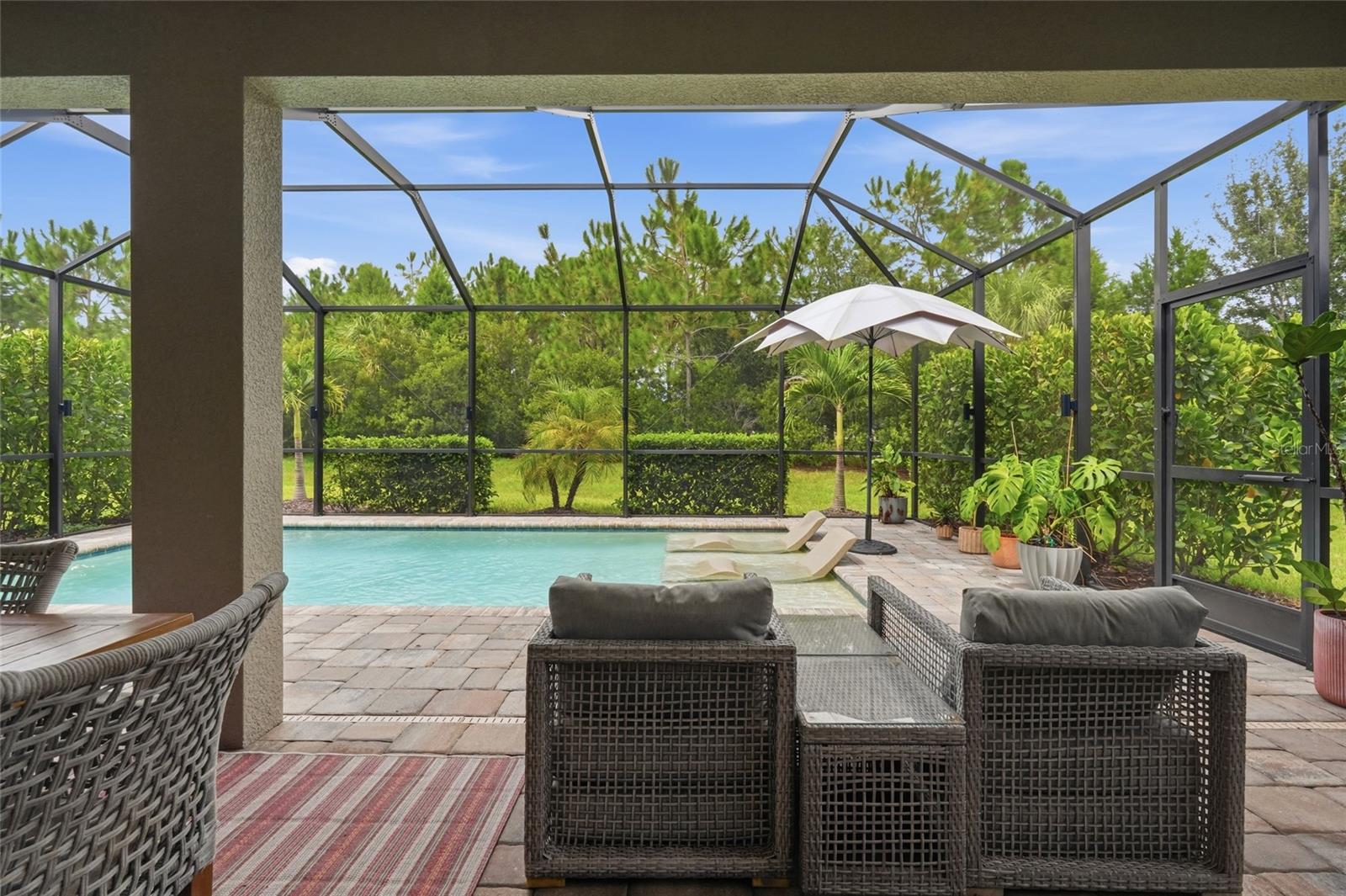 Screened pool patio with wicker lounge chairs, a white umbrella, and lush greenery.