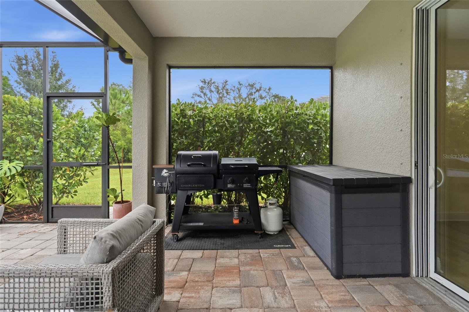 Screened patio with a wicker chair, black grill, paver flooring, and dark outdoor storage box.