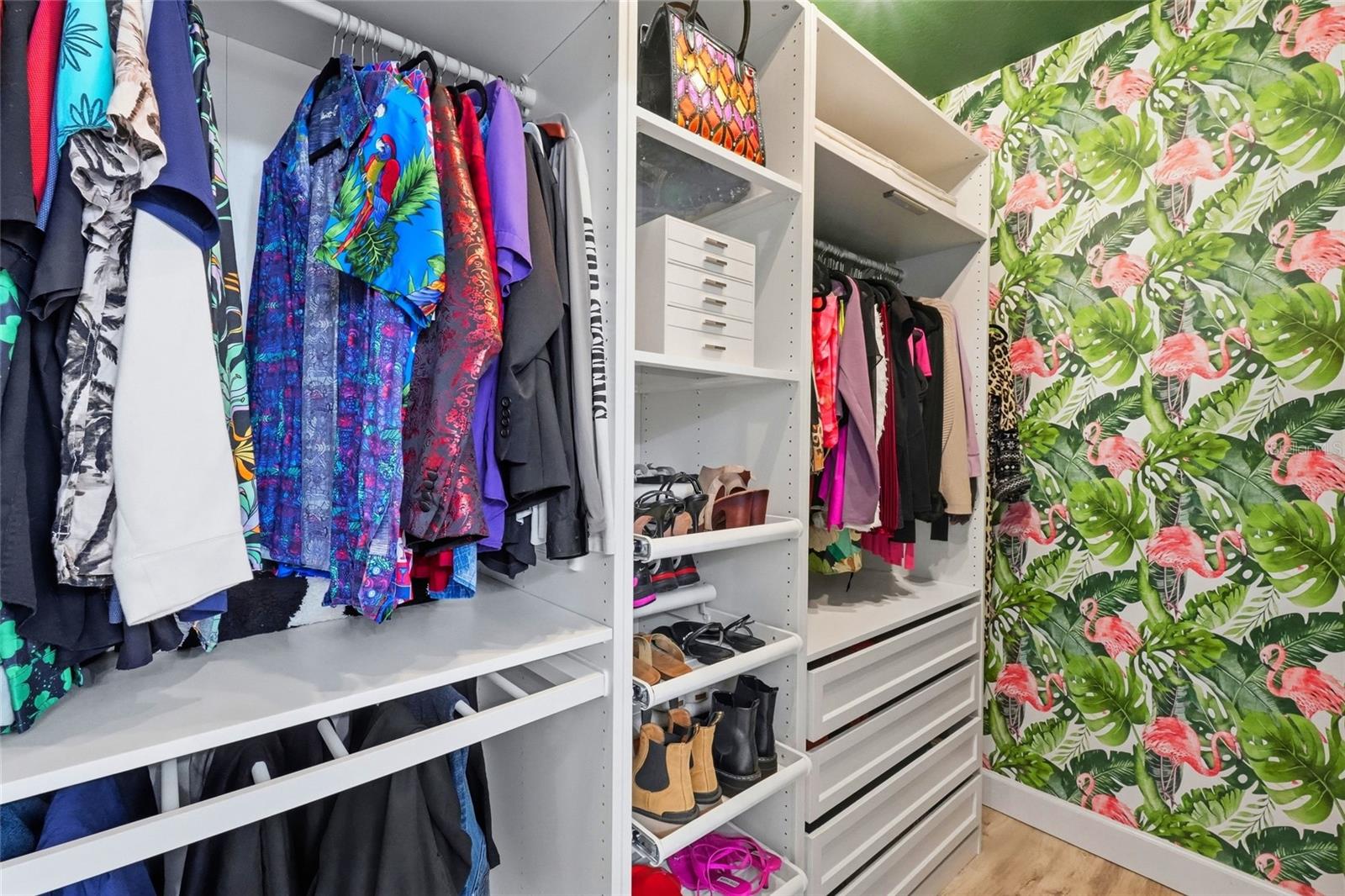 Walk-in closet with white built-ins, colorful clothes and shoes, and flamingo-and-foliage wallpaper.