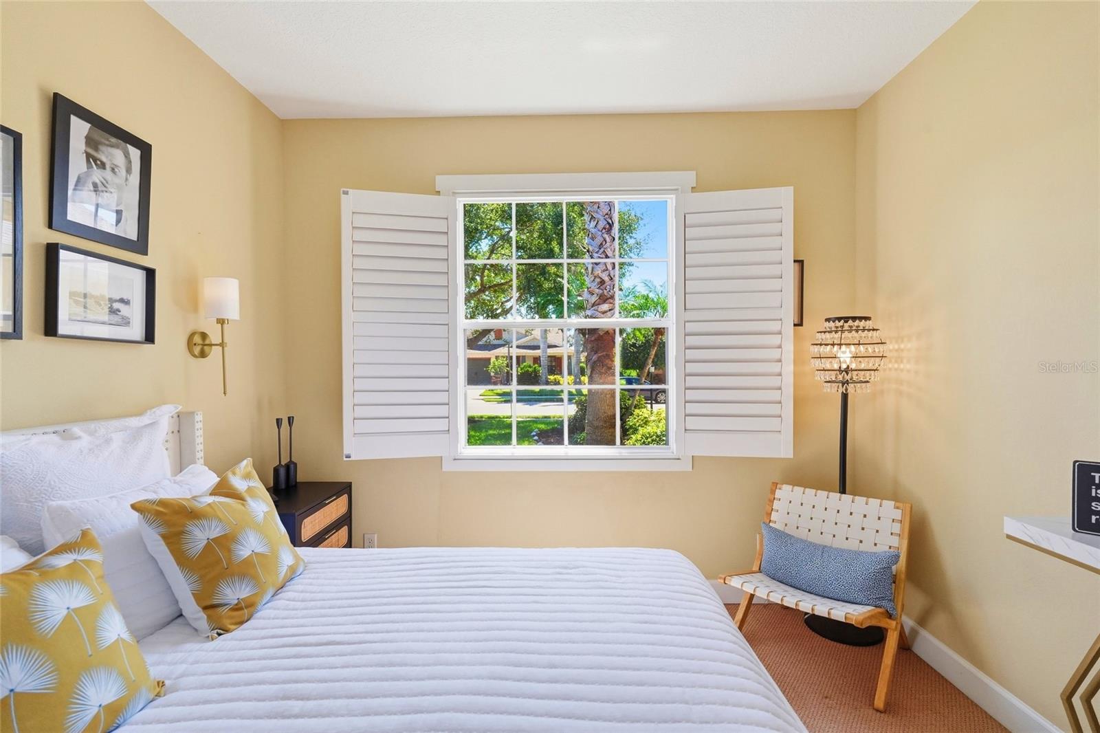 Bedroom with a white bed, yellow patterned pillows, and shuttered window overlooking palm trees.