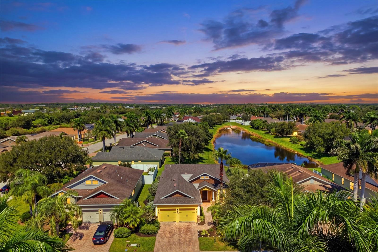 Aerial view of a palm-lined neighborhood and pond at sunset, with a home in the foreground.