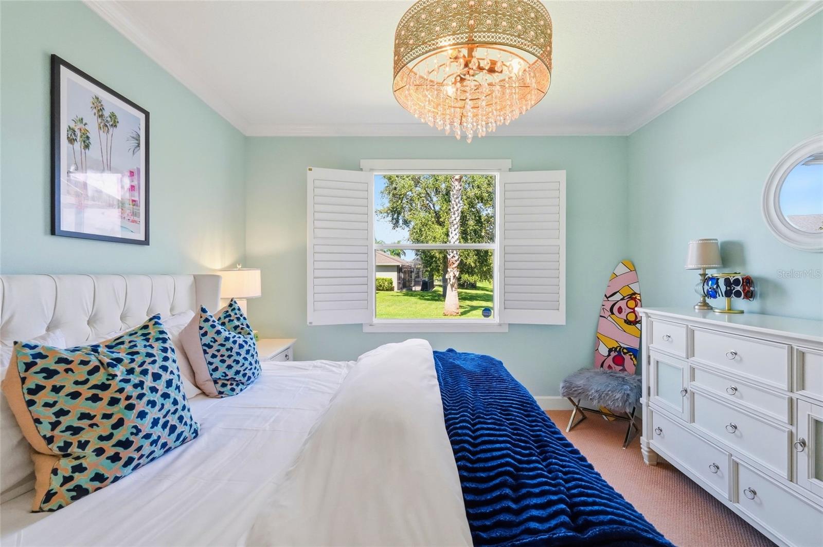 Bedroom with aqua walls, a white bed, colorful pillows, and open shutters overlooking a lawn and palm tree.