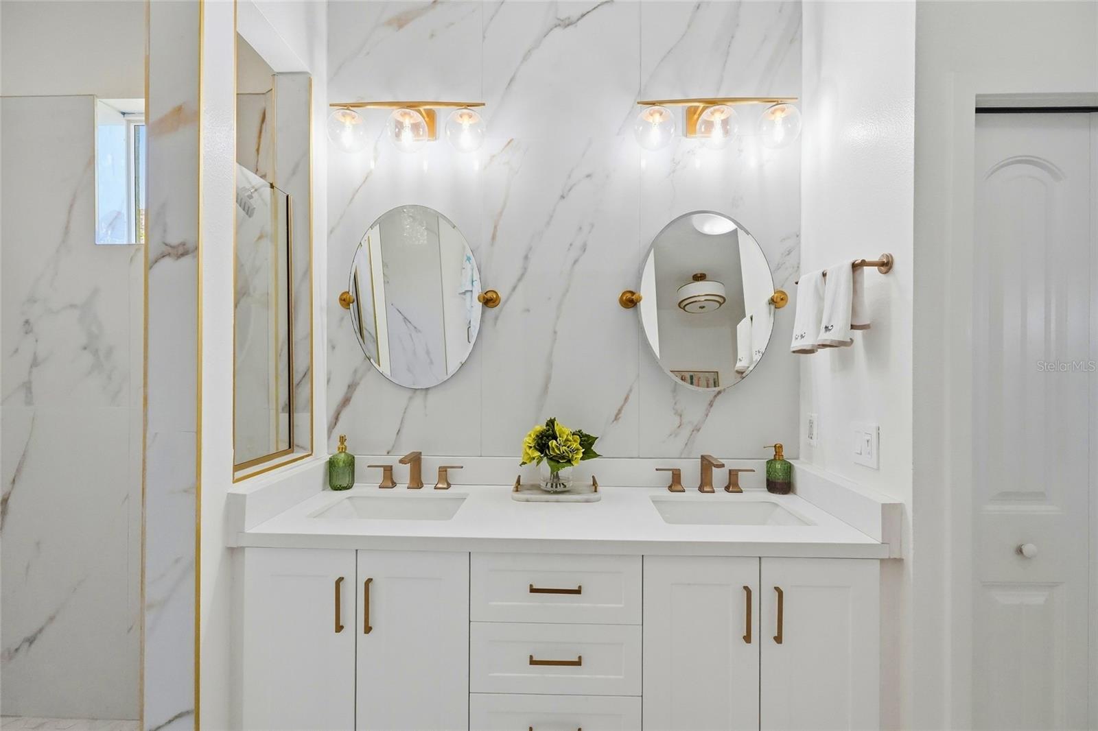 Bathroom with a double-sink white vanity, gold fixtures, oval mirrors, and marble-look tile.
