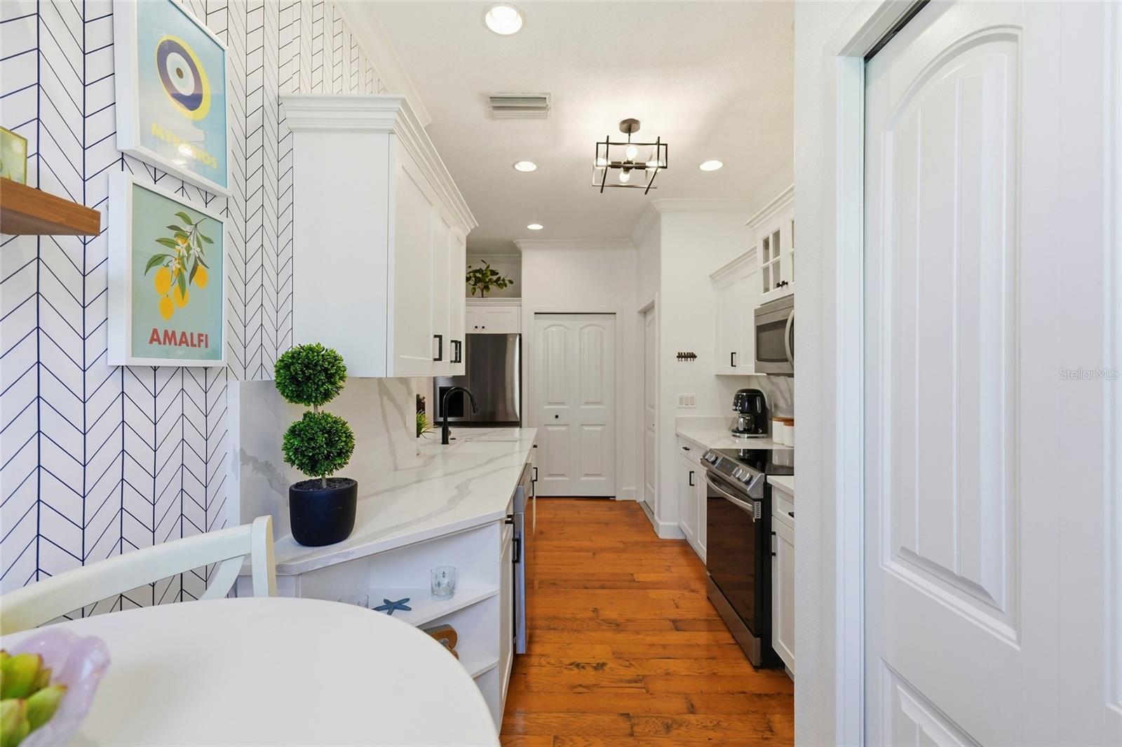 Bright galley kitchen with white cabinets, stainless appliances, warm wood floors, and a geometric accent wall.