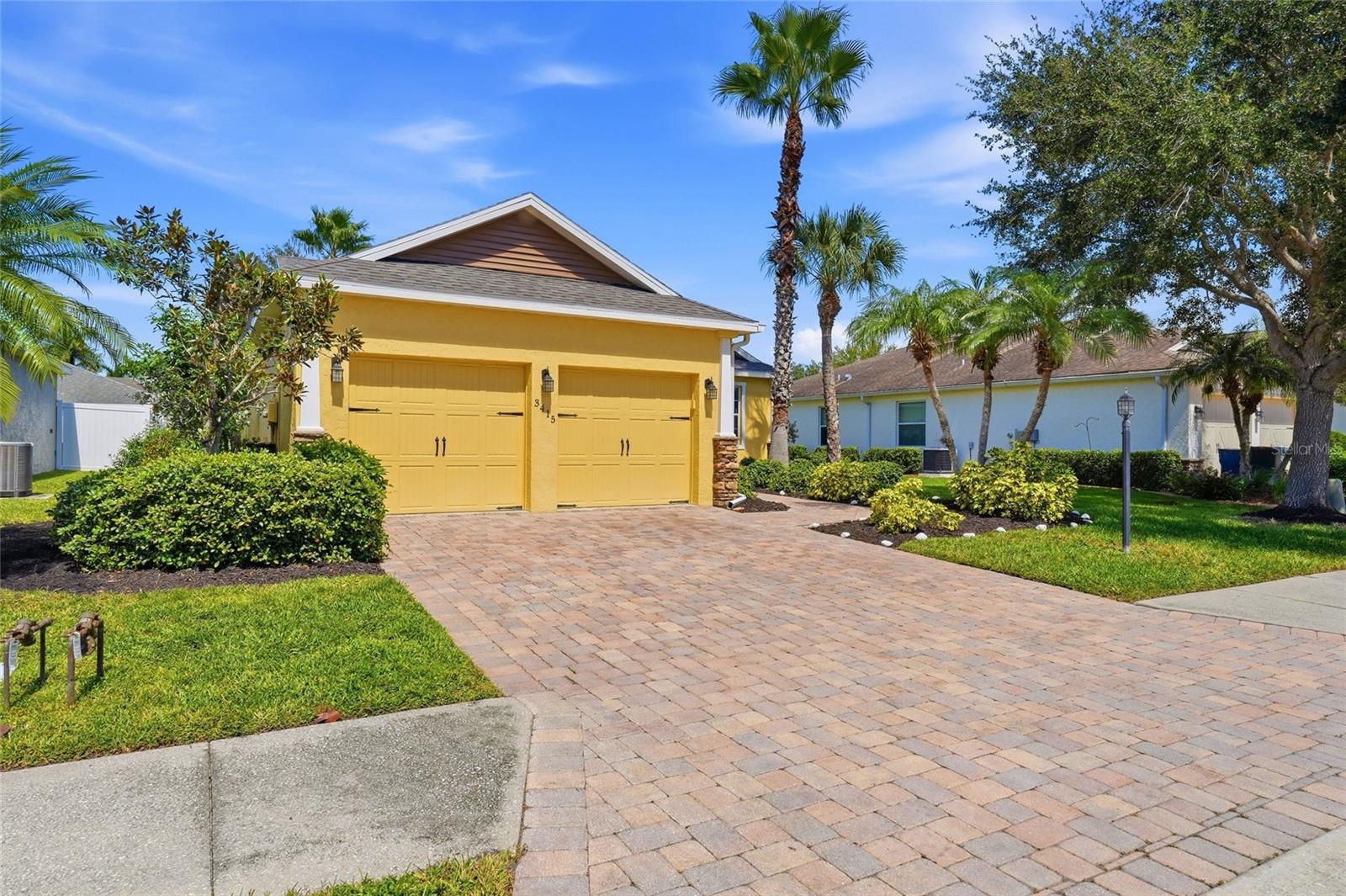 Yellow home with a double garage, brick-paver driveway, palms, and landscaped front yard.