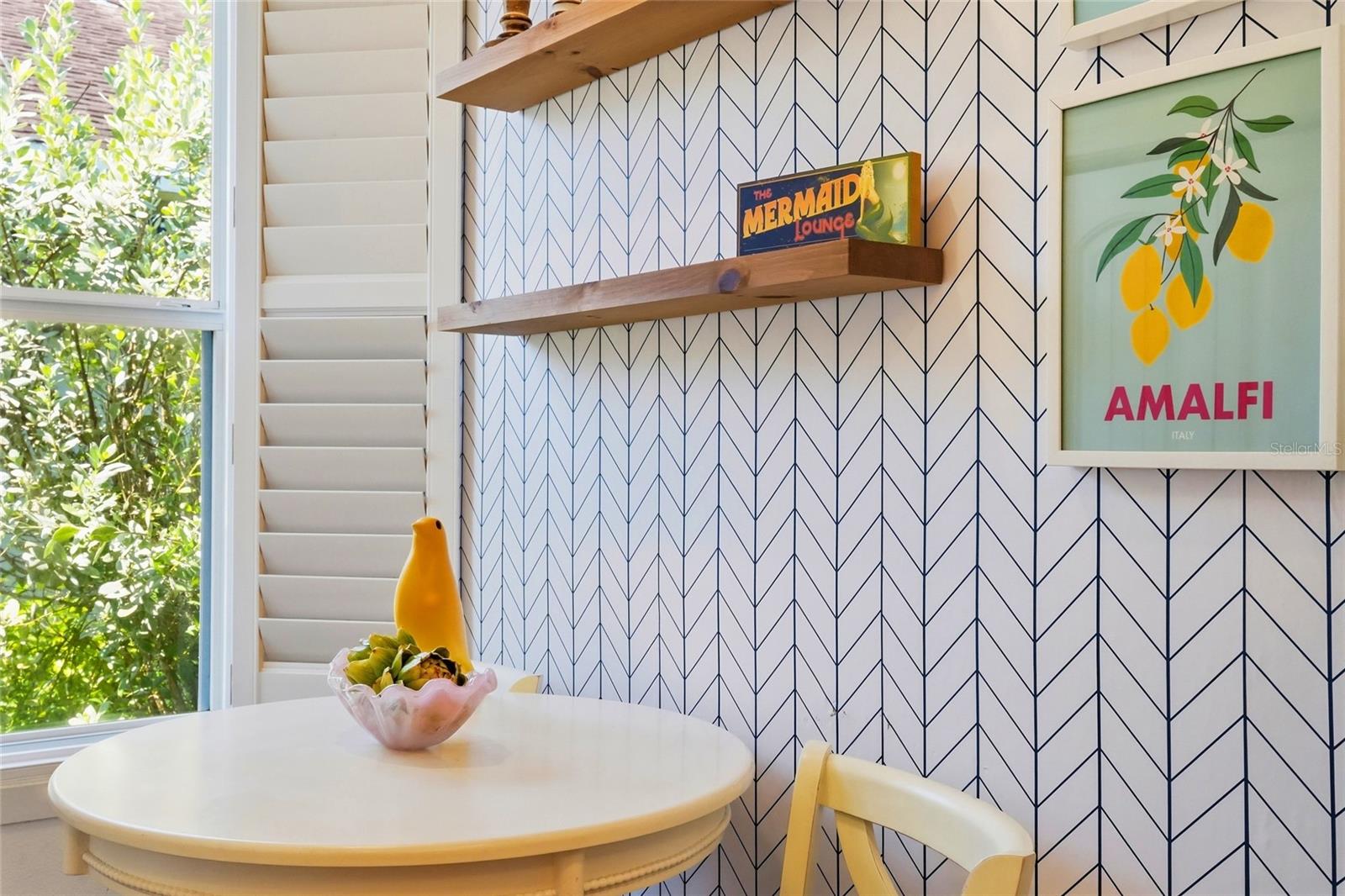 Dining nook with a round white table, patterned wall, wood shelves, and framed Amalfi lemon artwork.