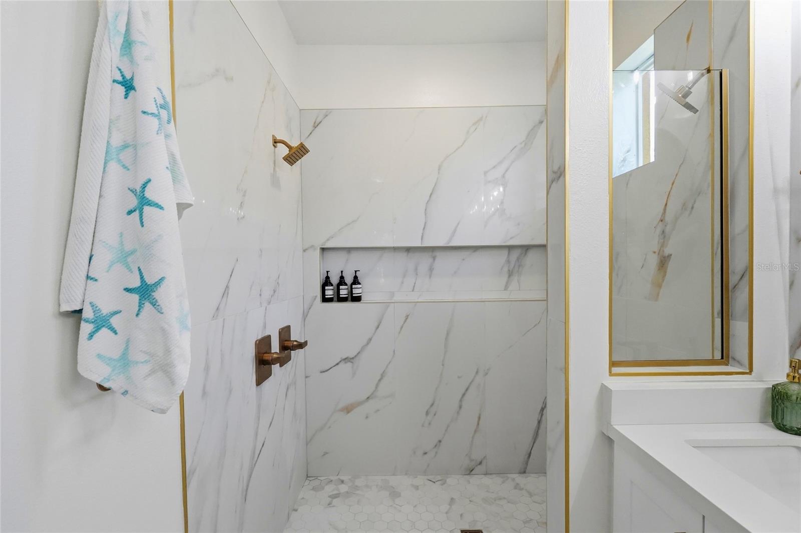 Bathroom with a marble-look walk-in shower, brass fixtures, gold-trimmed glass door, and starfish-patterned towel.