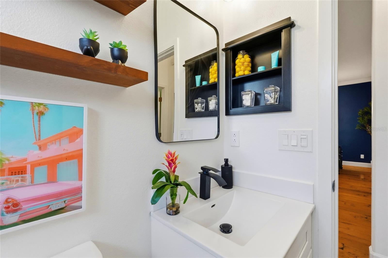 Bathroom vanity with a black faucet, large mirror, dark display shelves, and colorful framed artwork.
