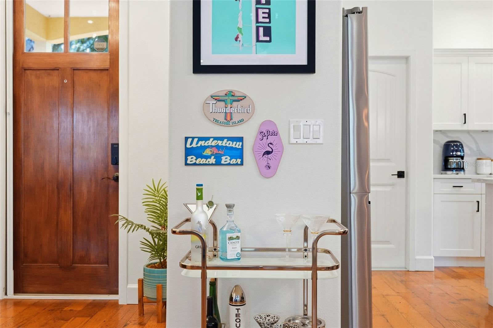 Entryway and kitchen with a wood front door, beach-themed wall signs, bar cart, and hardwood floors.
