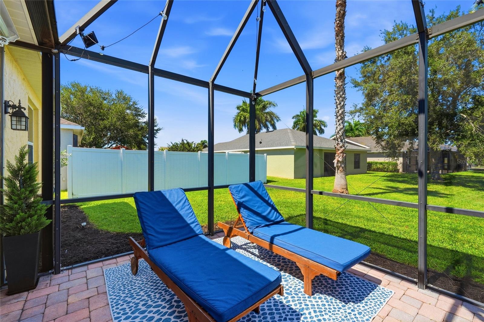 Screened lanai with two blue-cushioned loungers overlooking a fenced lawn and palm trees.